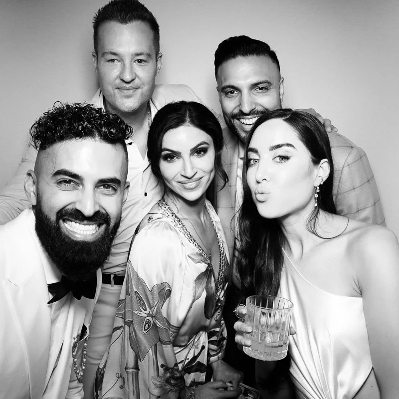 Group of five friends at a celebration, three men and two women, smiling and posing for a selfie, one woman holding a drink.