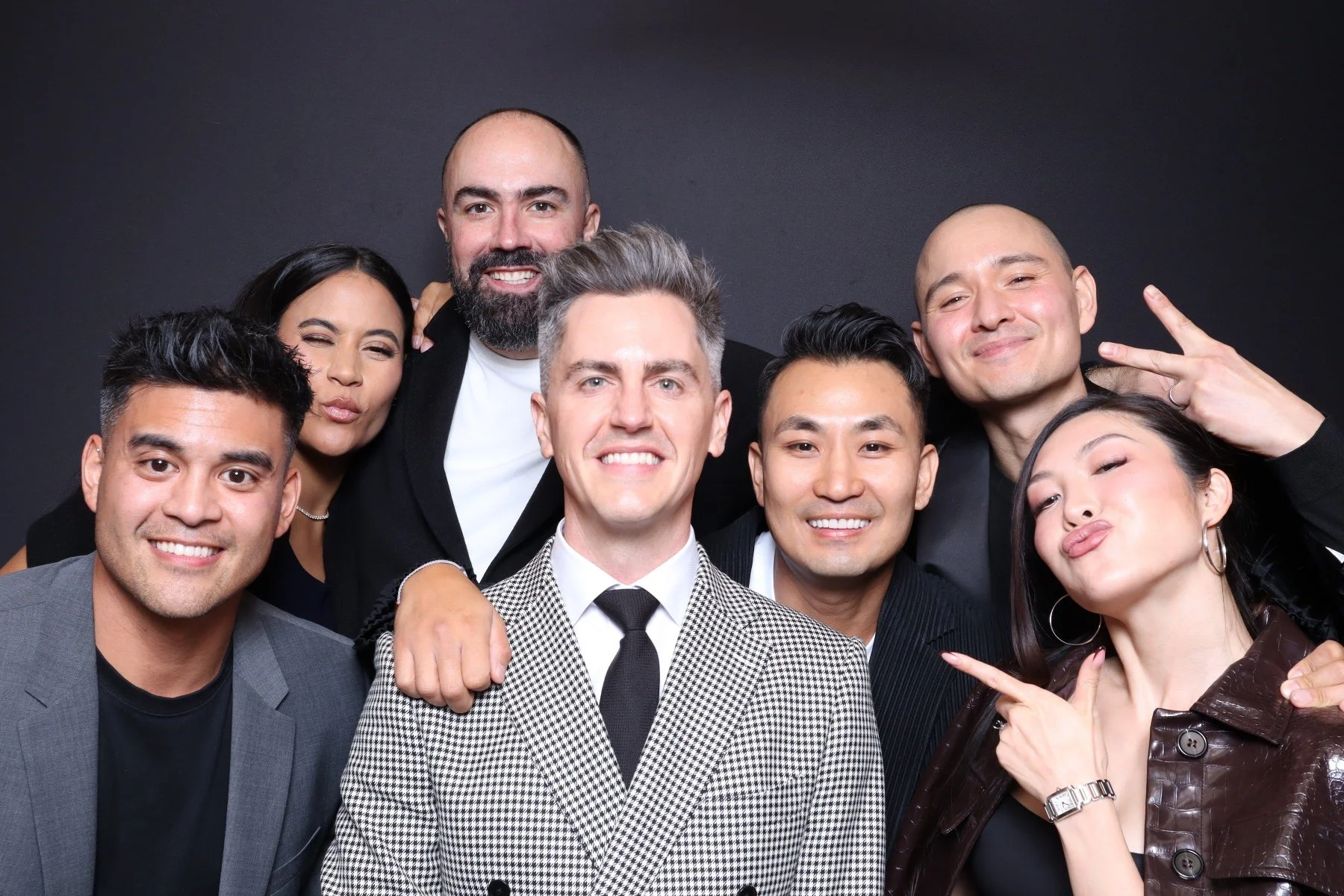 A diverse group of eight people posing together against a black background, smiling and making playful gestures.