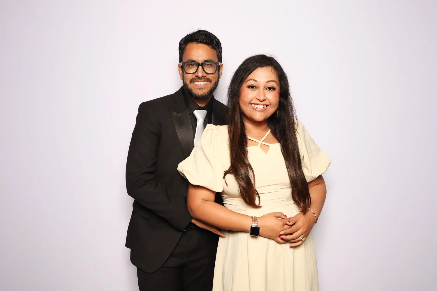A smiling couple, man in a black suit and glasses, woman in a light yellow dress, standing against a white background.
