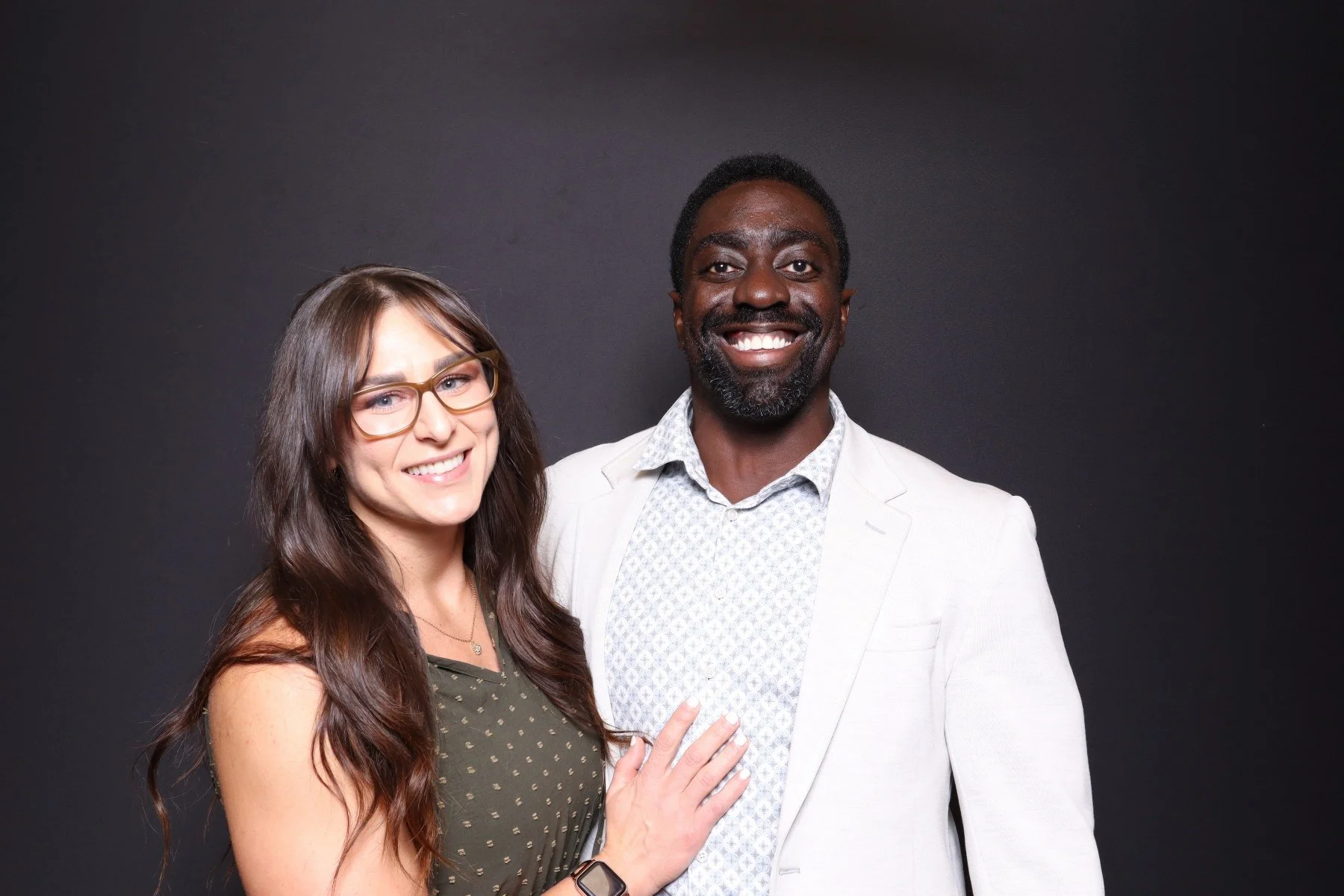 A smiling woman with long brown hair, glasses, wearing a green patterned sleeveless top, and a man with short black hair, beard, wearing a white blazer over a patterned shirt, standing against a black background.