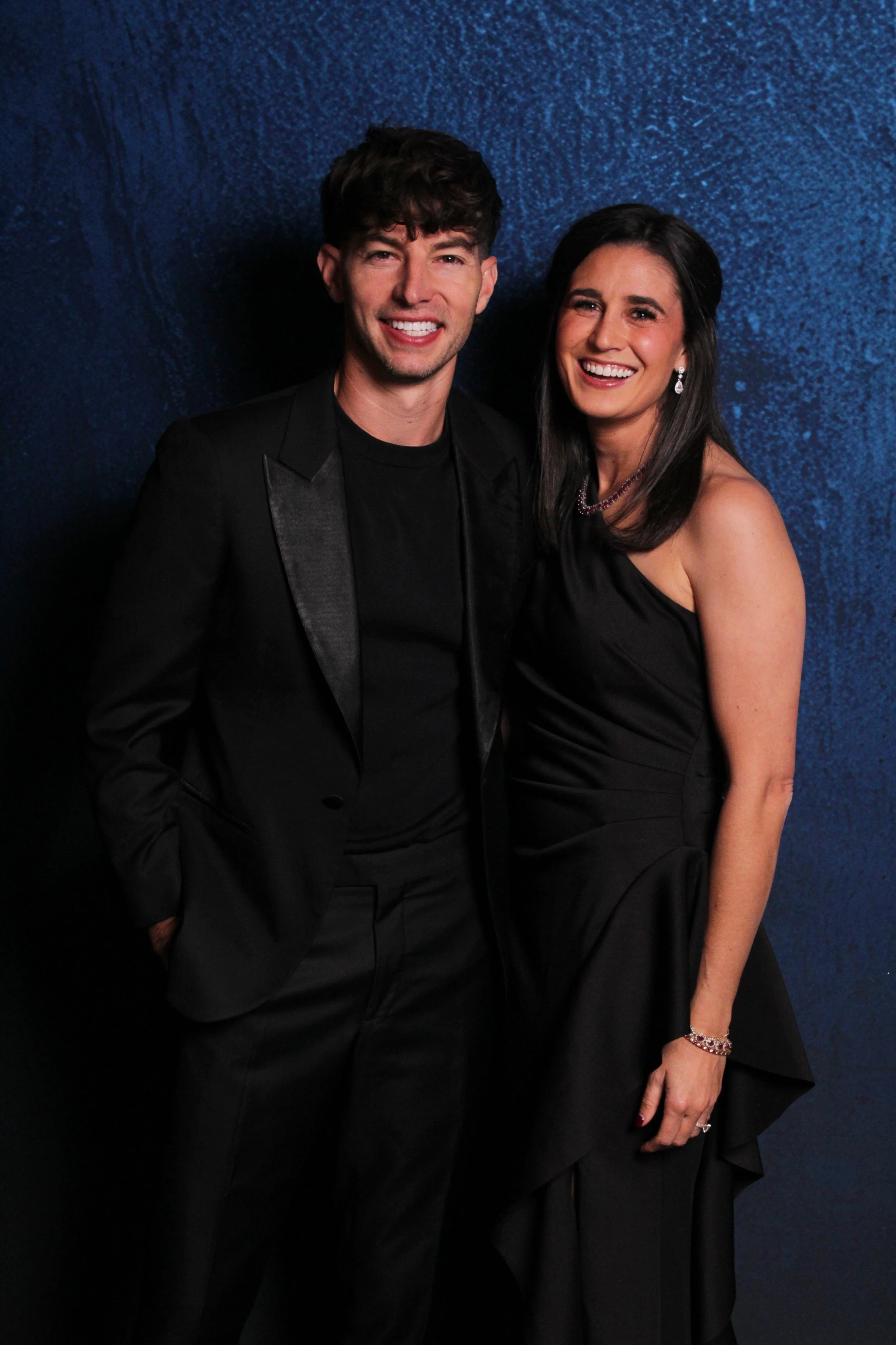 A man and a woman dressed in formal black attire, smiling at the camera, standing in front of a blue textured backdrop.