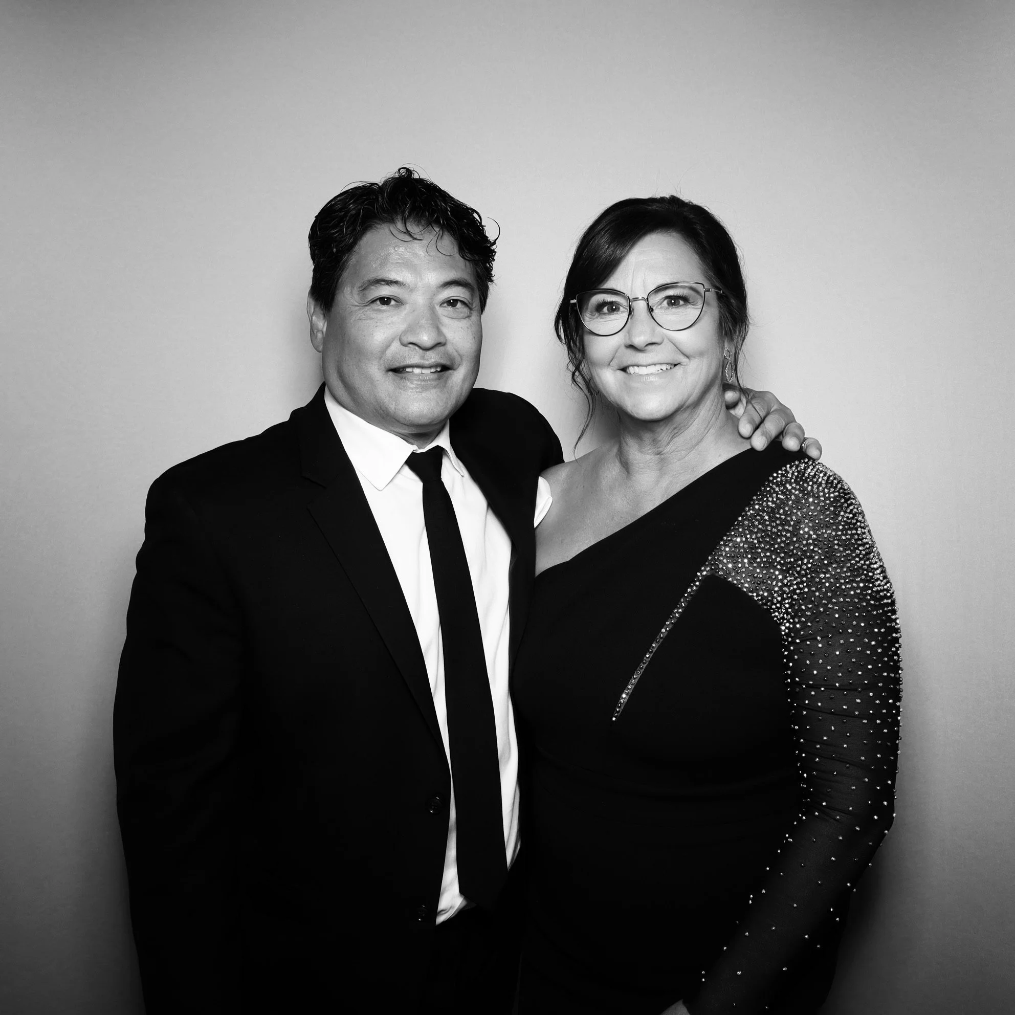 Black-and-white photo of a man in a suit and a woman in a dress with glasses, smiling and standing close together, the man has his arm around the woman's shoulder.
