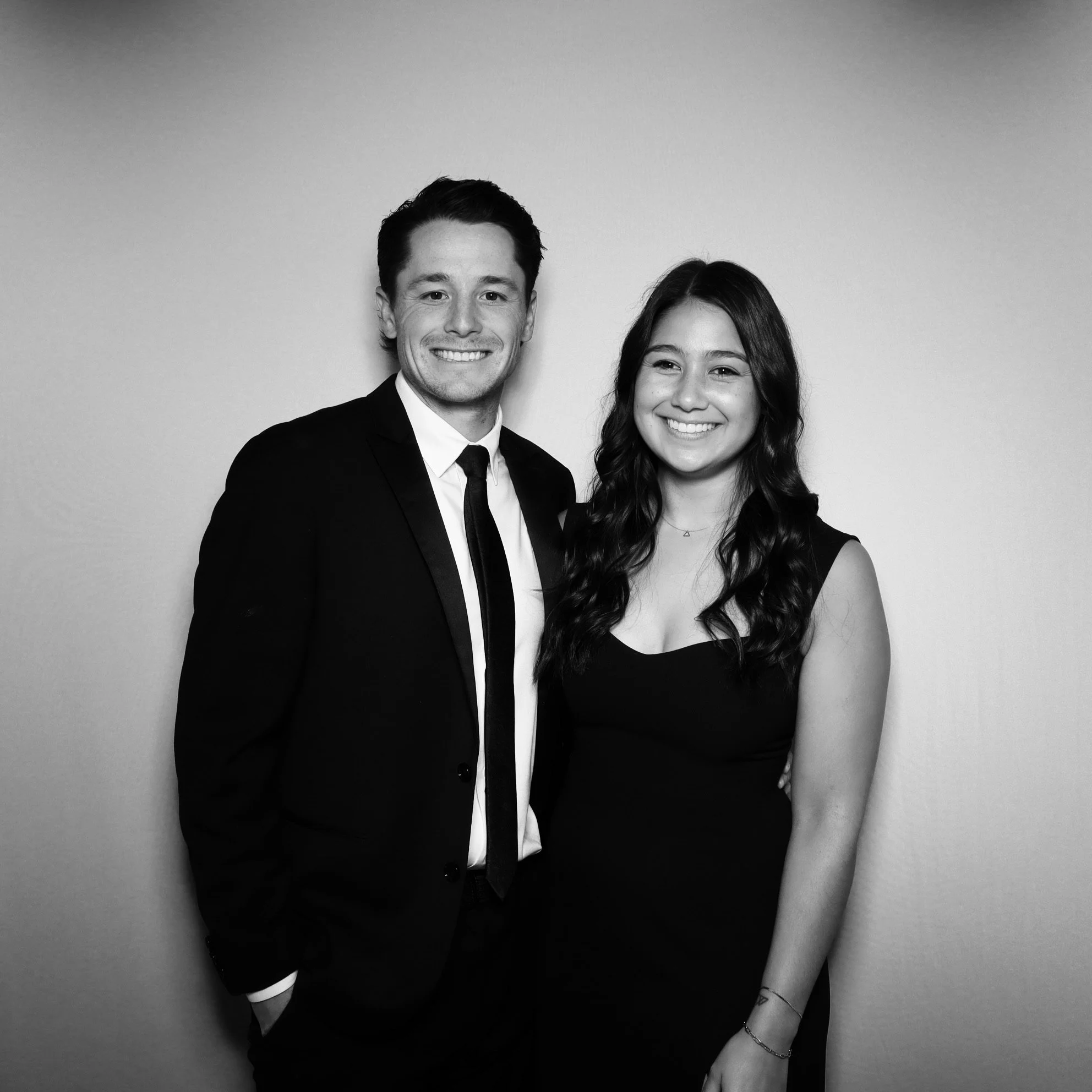 Black-and-white photo of a young man and woman smiling and standing together against a plain background; the man is wearing a suit and tie, and the woman is in a sleeveless dress.