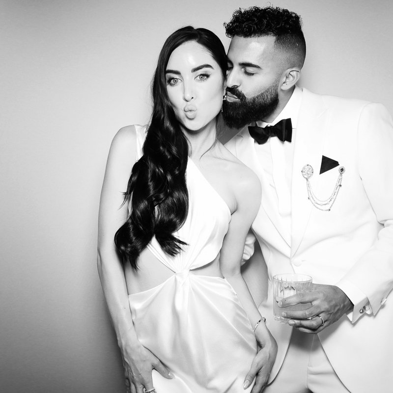 A woman with long dark hair making a kissy face next to a man in a tuxedo with a bow tie, who is whispering in her ear. The woman is wearing a white dress with a cutout, and the man is holding a glass.