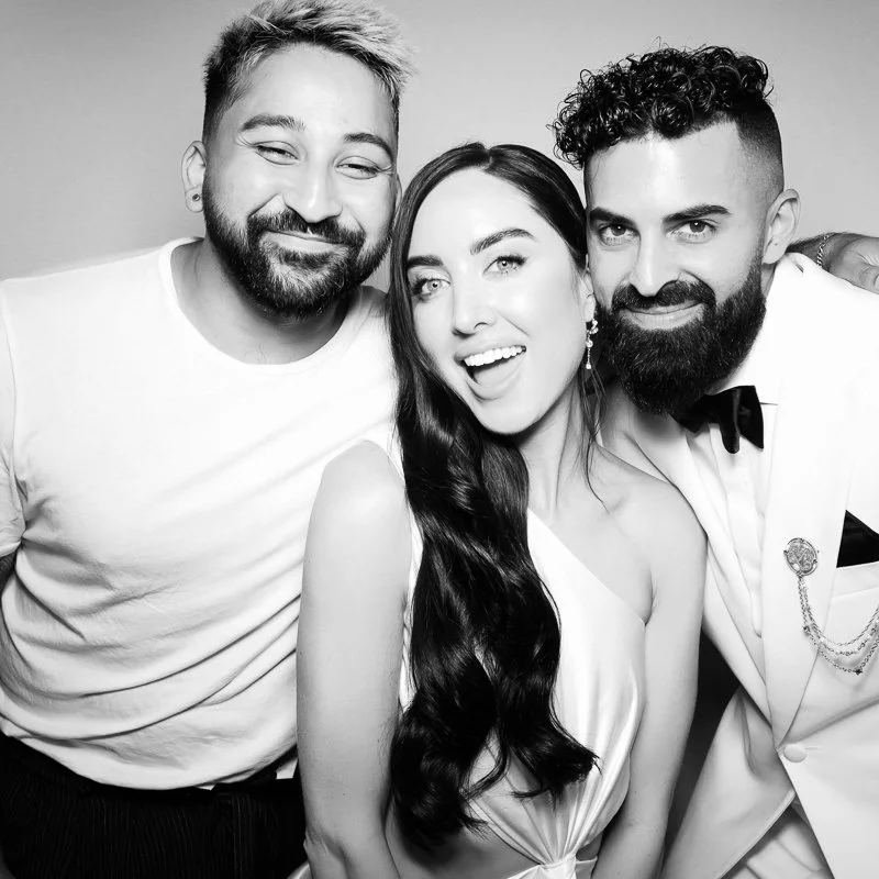 Three smiling people in formal attire taking a group selfie in black and white.