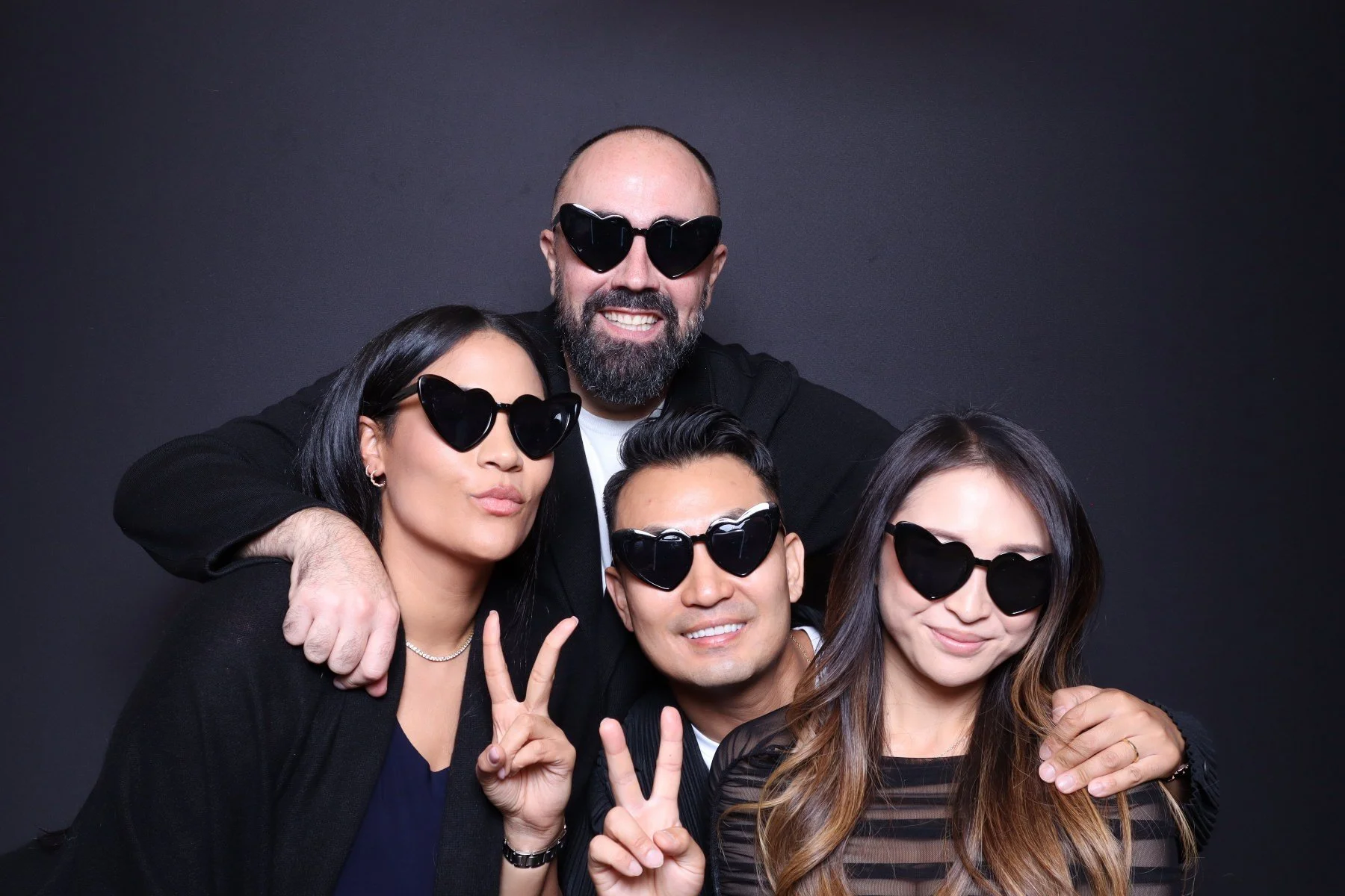 Group of four friends wearing black sunglasses, posing together against a black background. They are smiling and making peace signs.