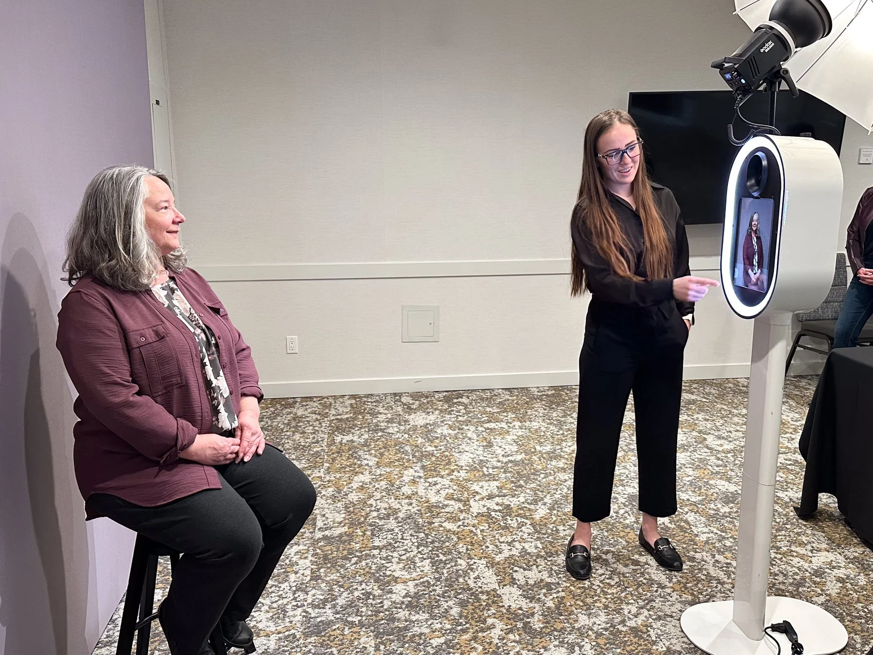 A woman with gray hair sitting on a stool, a young woman with long brown hair standing in front of a large machine with a screen showing her portrait, in a room with a carpeted floor and plain walls.