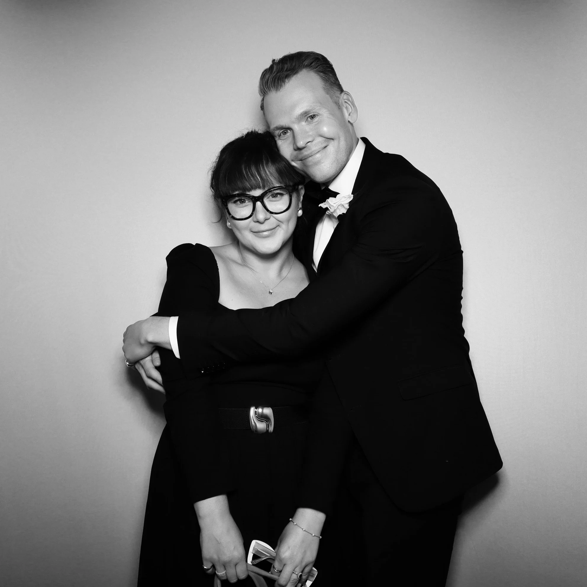 A black and white photo of a smiling man in a tuxedo hugging a woman with glasses and a dress, both looking at the camera.