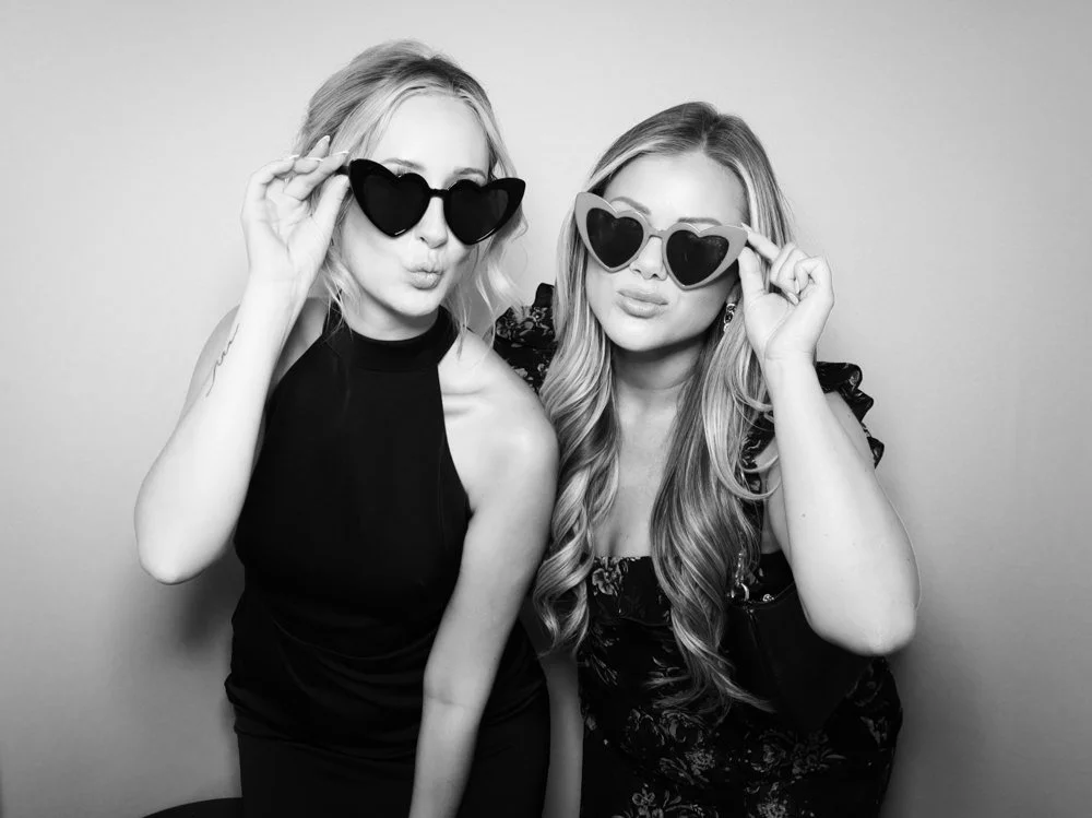 Two young women with light hair wearing black and patterned outfits, holding heart-shaped sunglasses near their eyes, posing playfully against a plain background.