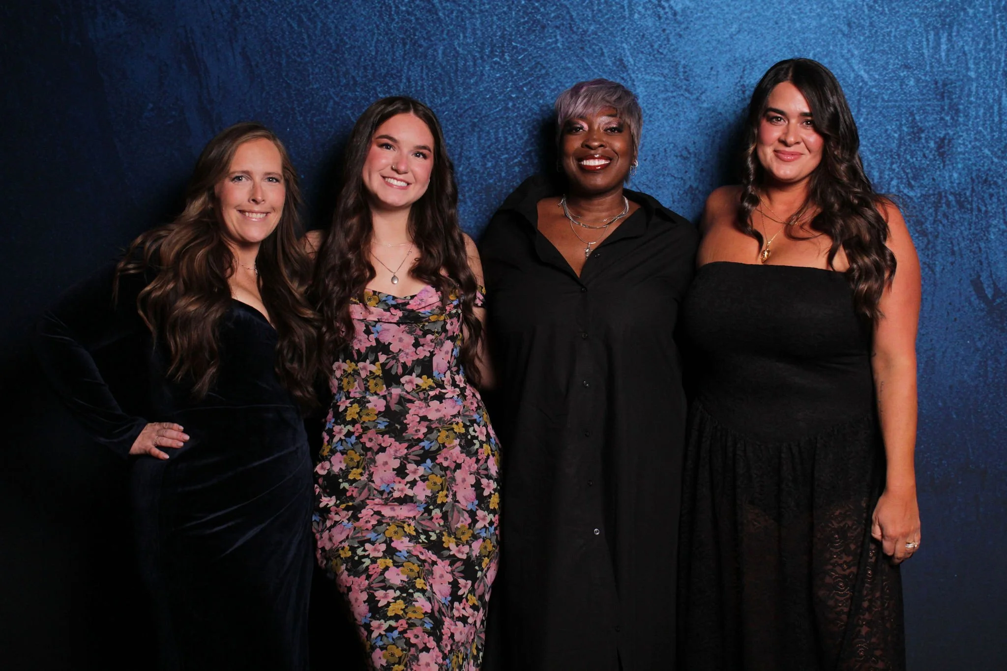 Four women standing together against a dark blue textured background, smiling and dressed in black and floral clothing