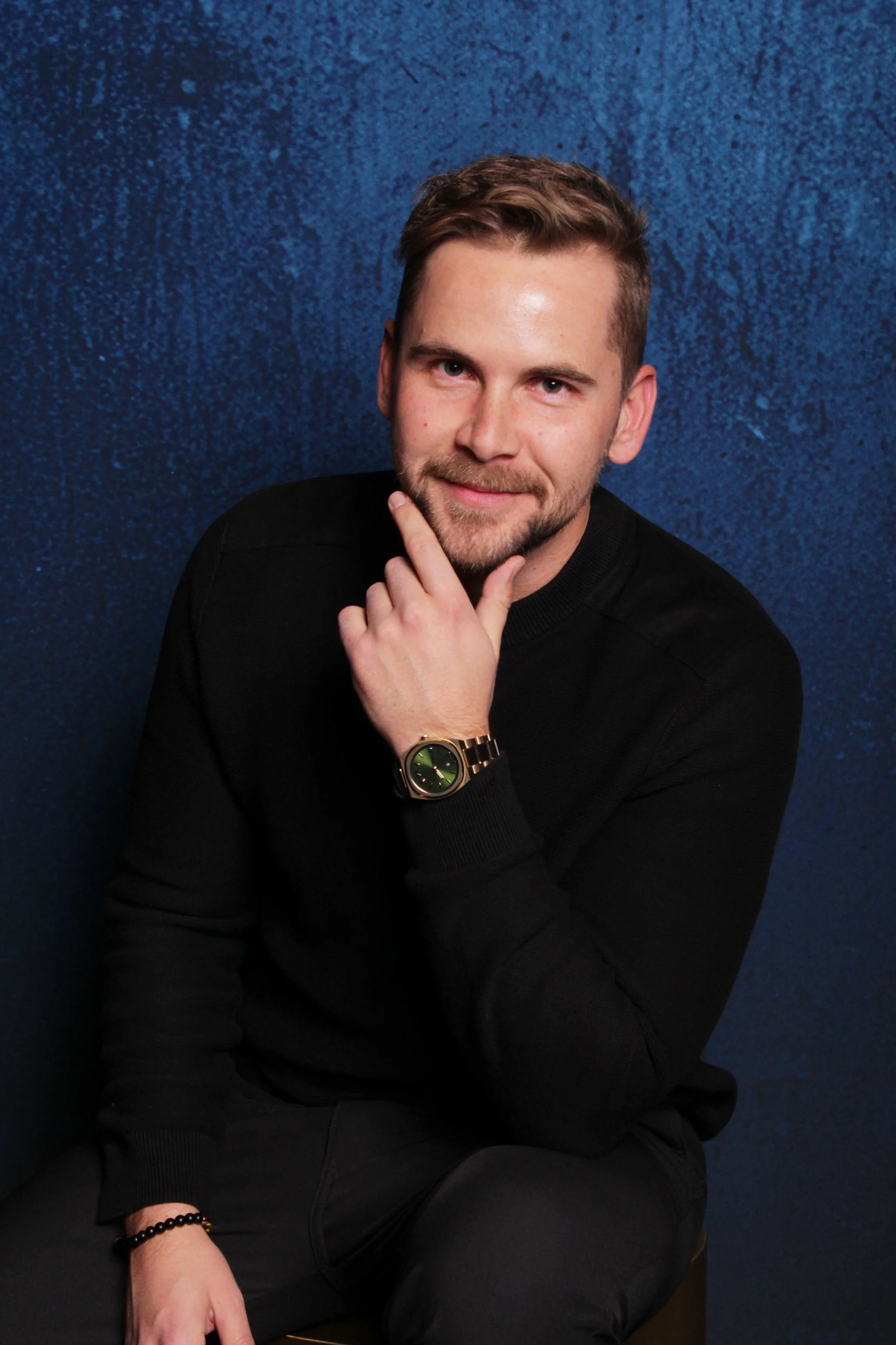 A young man with light brown hair and a beard, wearing a black sweater and a gold watch, sitting against a dark blue textured background, smiling and resting his chin on his hand.
