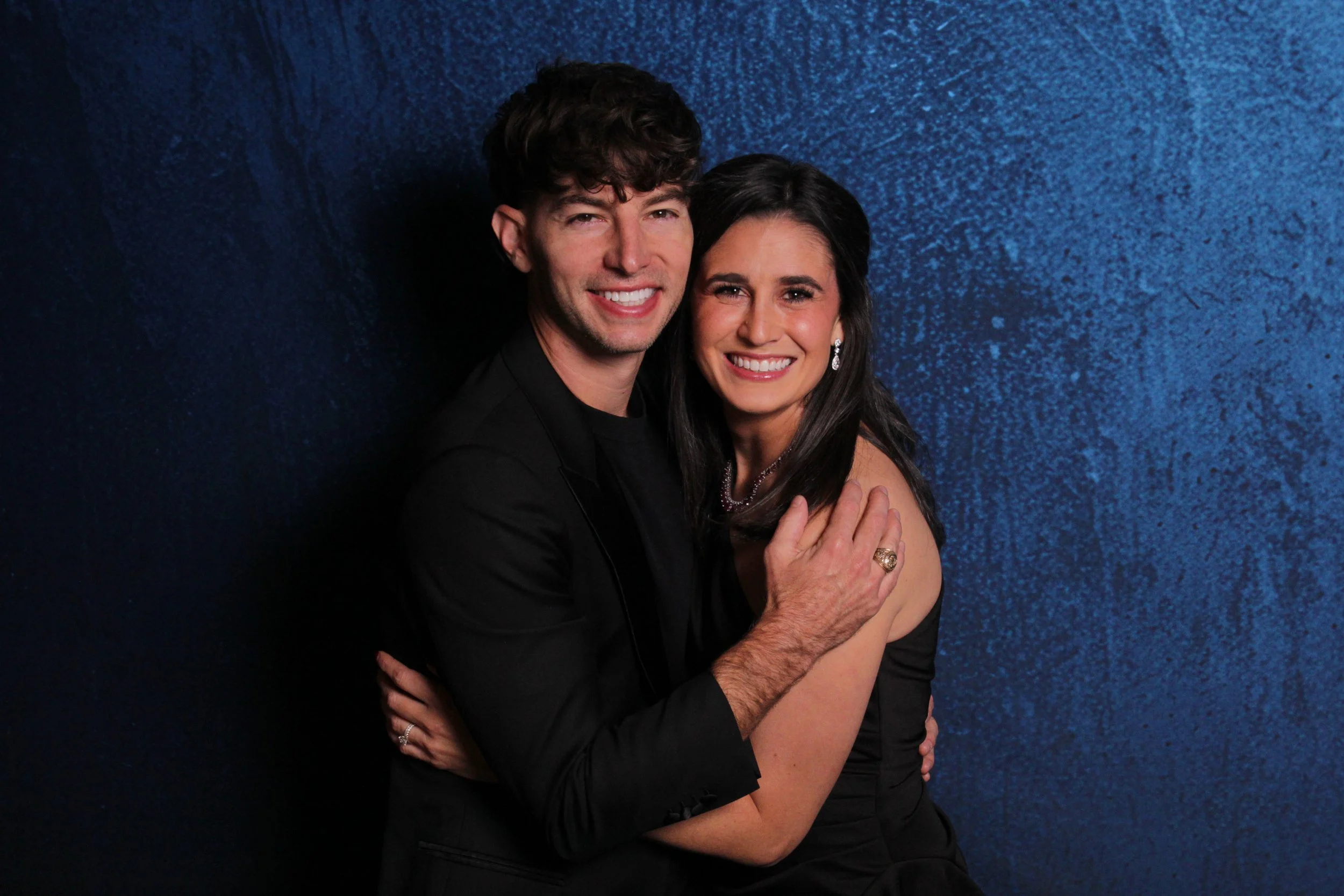 A smiling man and woman in formal black attire embracing each other against a blue textured background.