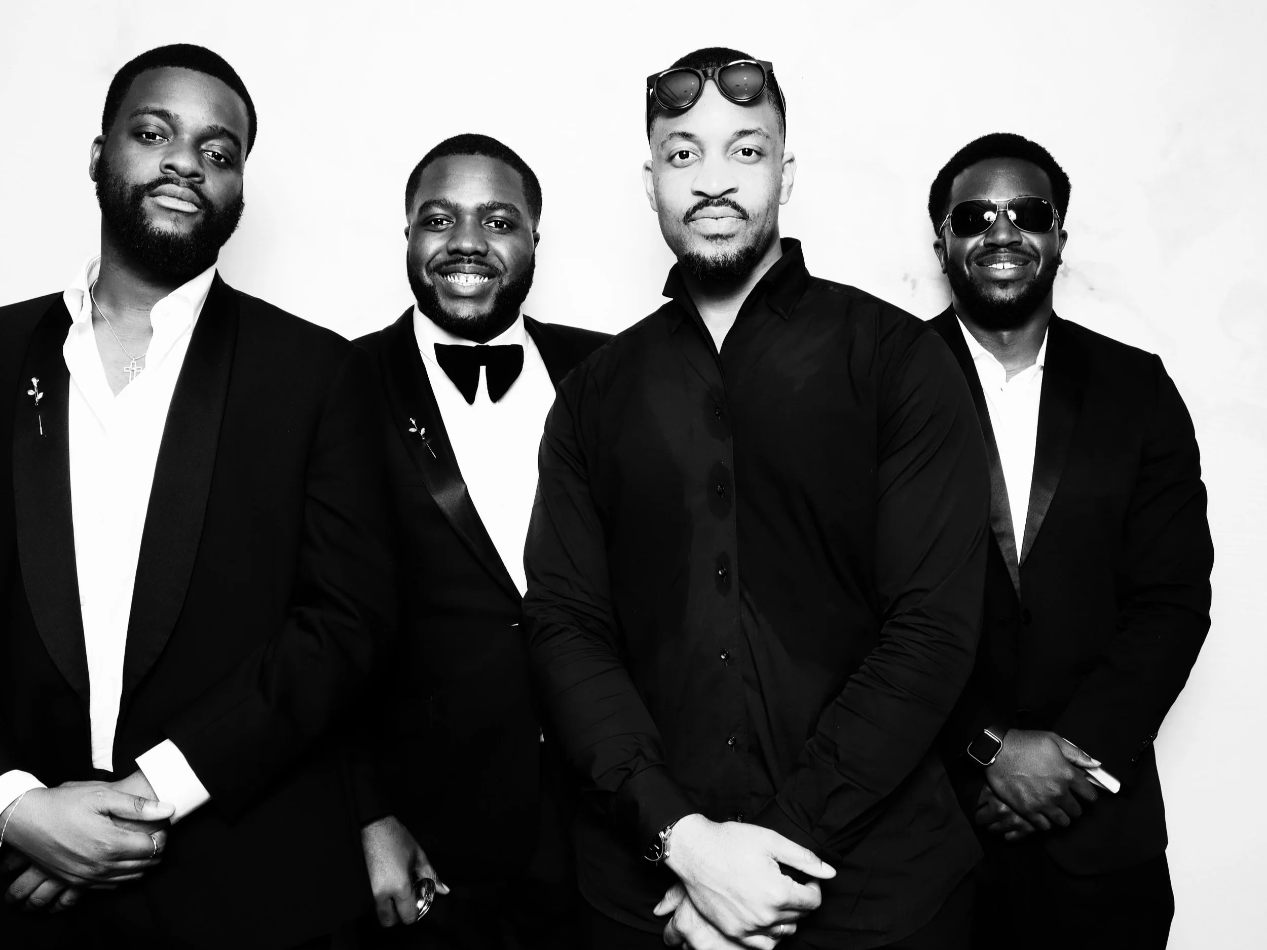 A black-and-white photo of five men dressed in formal suits and tuxedos, with two wearing sunglasses, standing against a plain background.