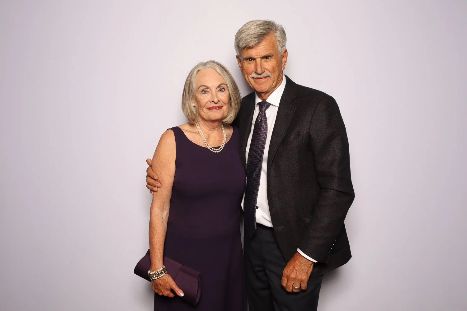 An elderly couple, a woman with short gray hair wearing a purple dress with pearl necklace, and a man with gray hair wearing a black suit with white shirt and tie, standing close together against a plain light gray background.