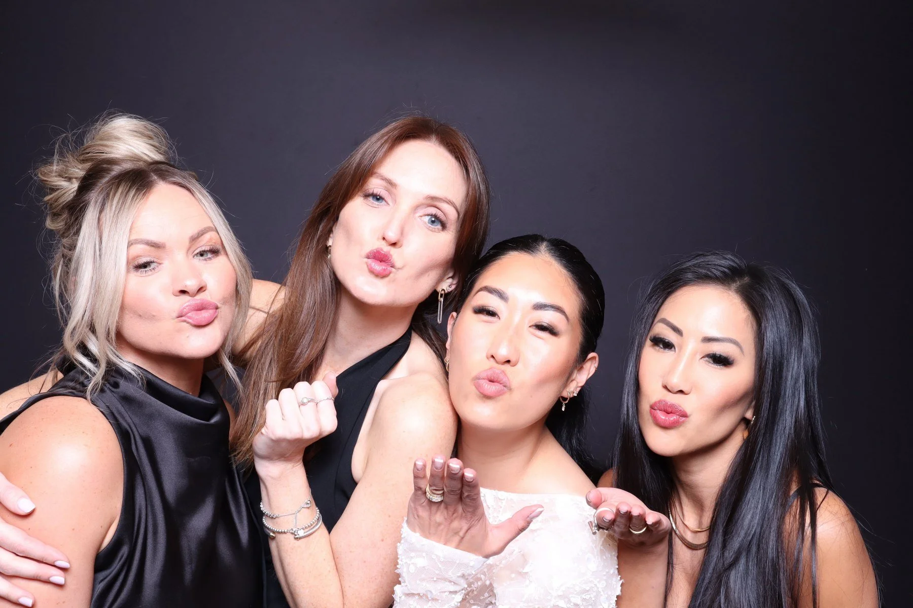 Four women posing together, making kissing faces at the camera, against a dark background.