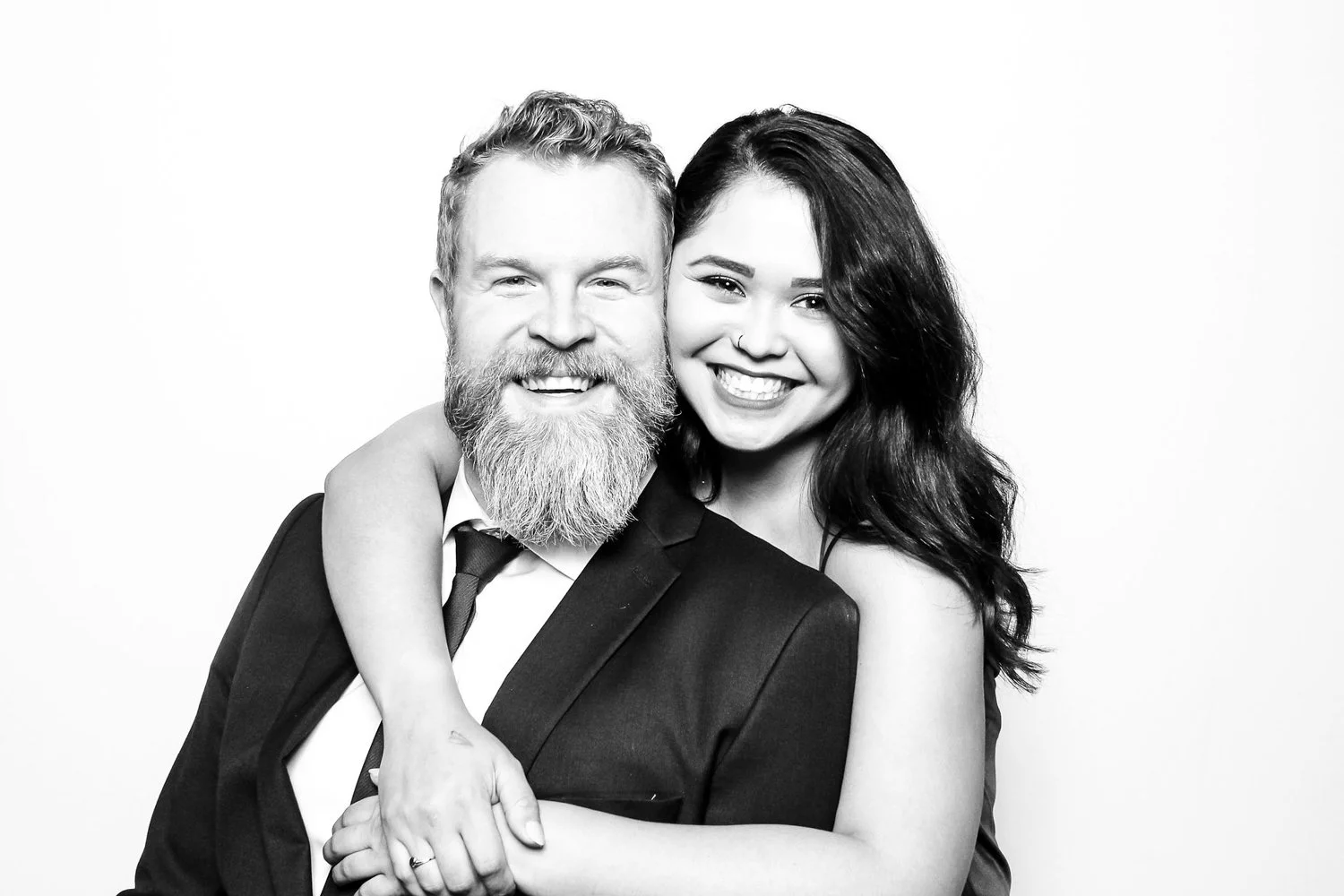 A smiling bearded man in a suit with a woman embracing him, both facing the camera against a plain background.