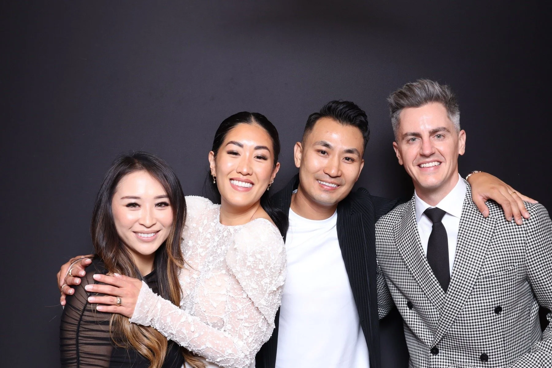 Group of four happy diverse people smiling and posing together against a black background, with arms around each other.