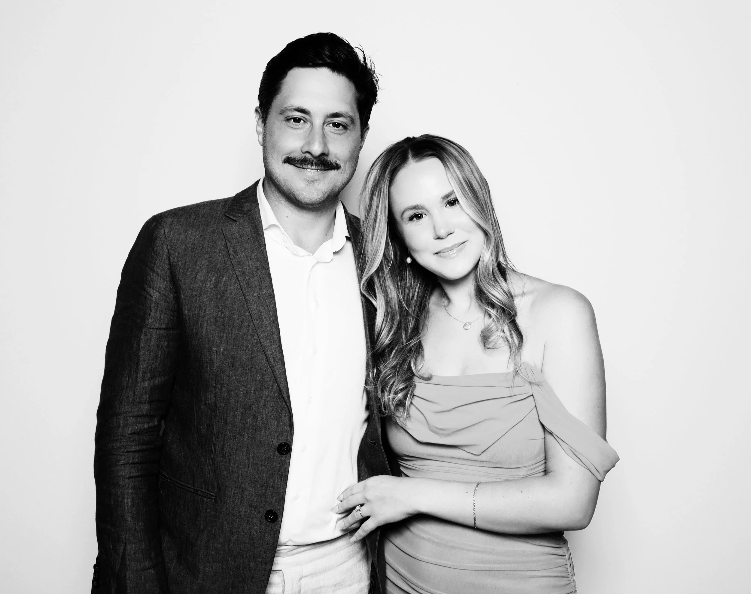 A black and white photograph of a man and woman standing close together, smiling at the camera. The man is wearing a suit jacket and white shirt, and the woman is in a strapless dress with wavy hair.