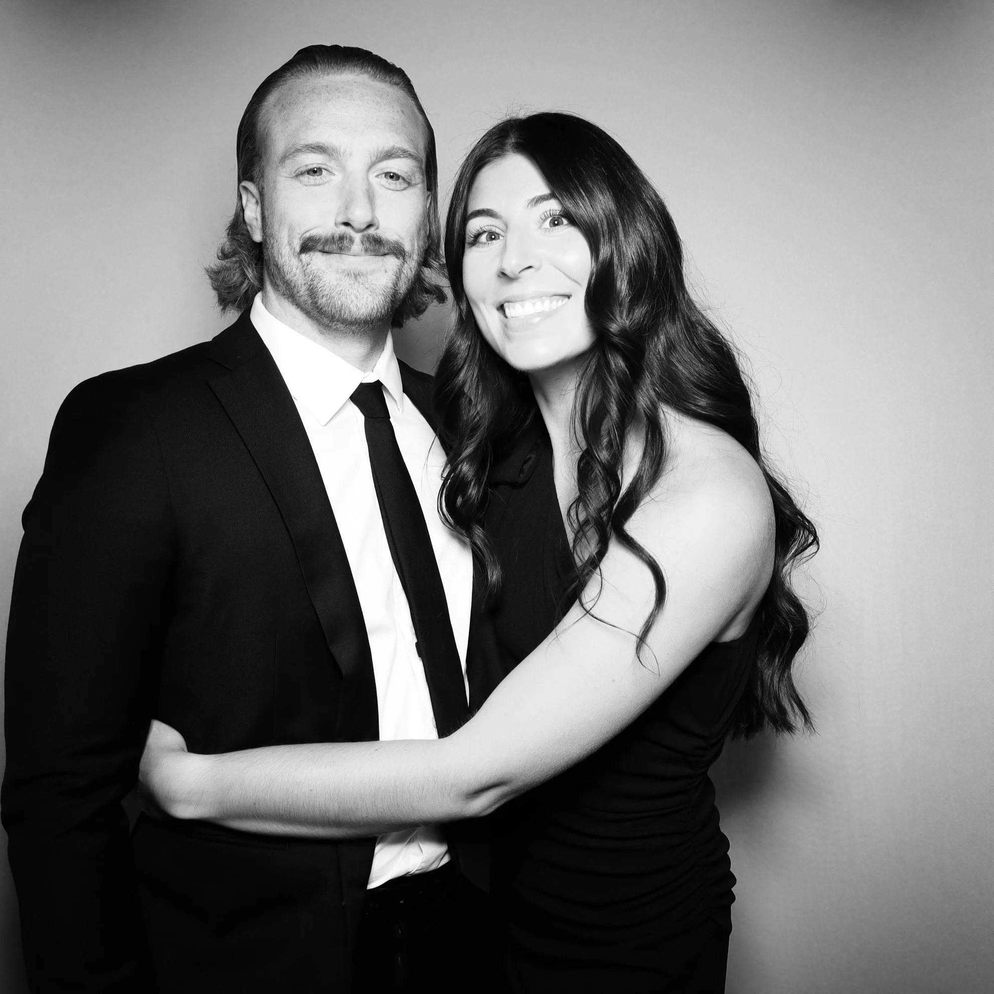 Black and white photo of a smiling man in a suit and tie and a woman with long wavy hair wearing a sleeveless dress, embracing each other, against a plain background.