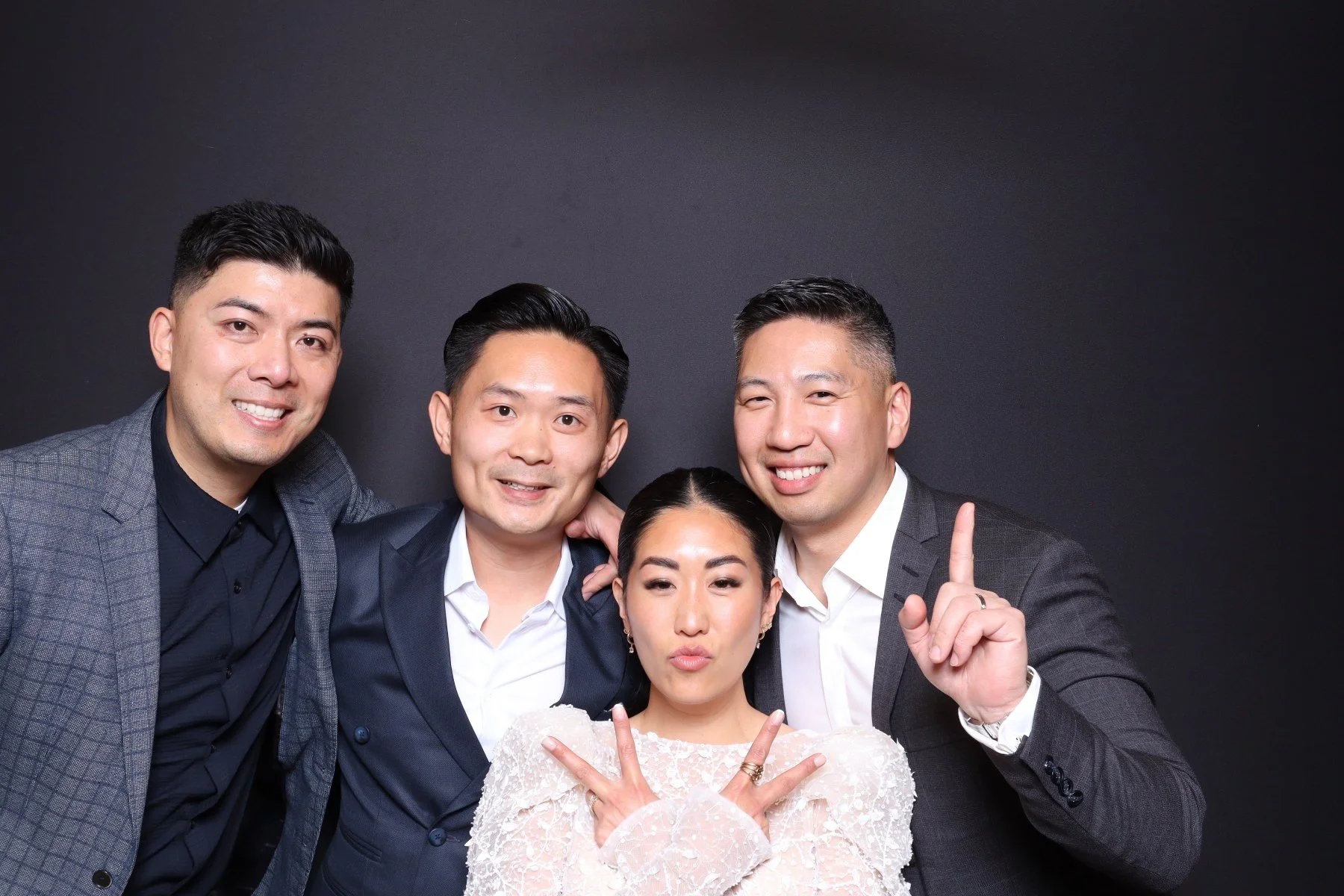 Four people, three men and one woman, posing together against a dark background. They are smiling and making gestures, with the woman in the center making a peace sign with both hands. All are dressed in formal or semi-formal attire.