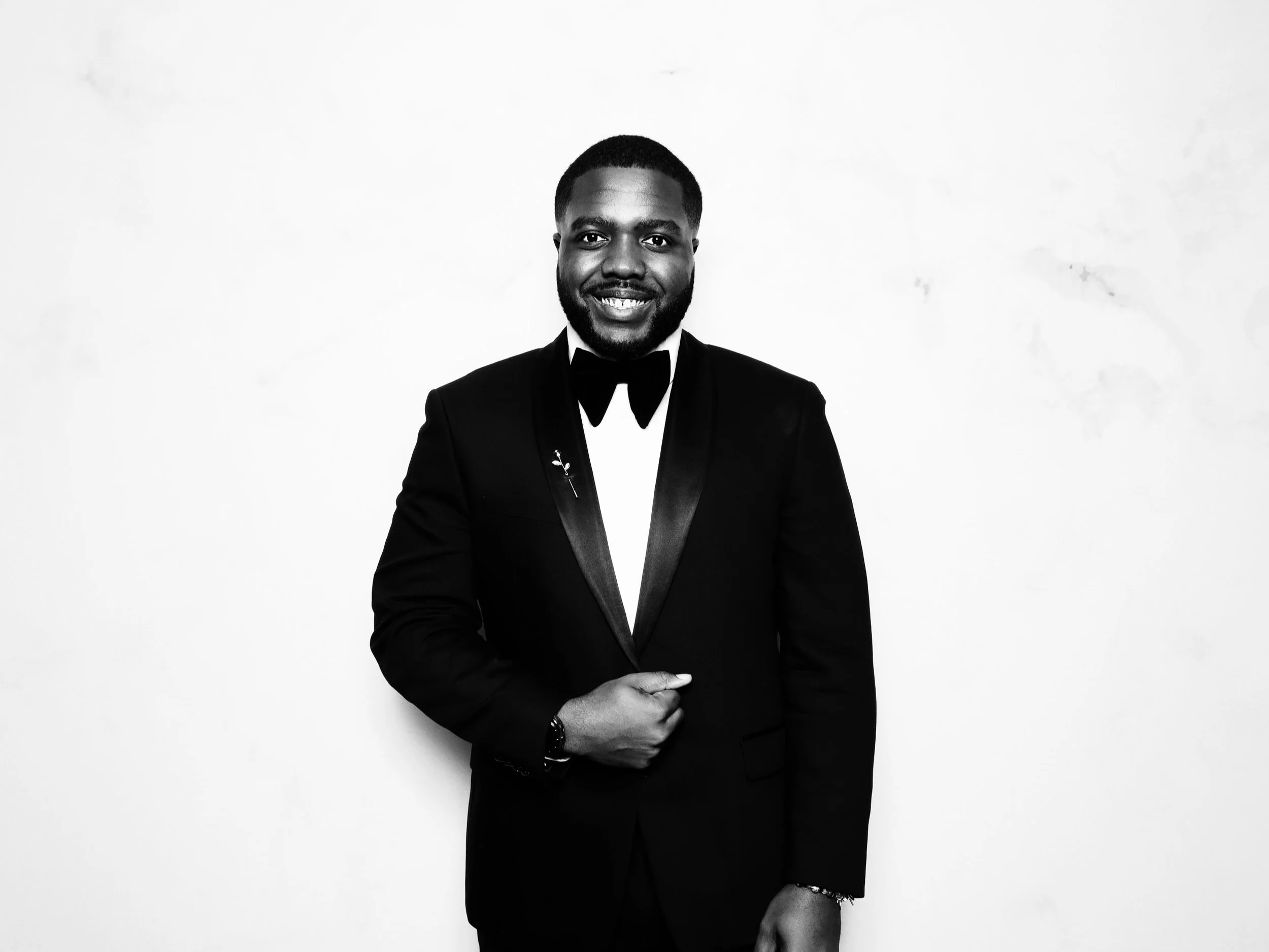 A smiling man in a tuxedo standing against a plain white background.