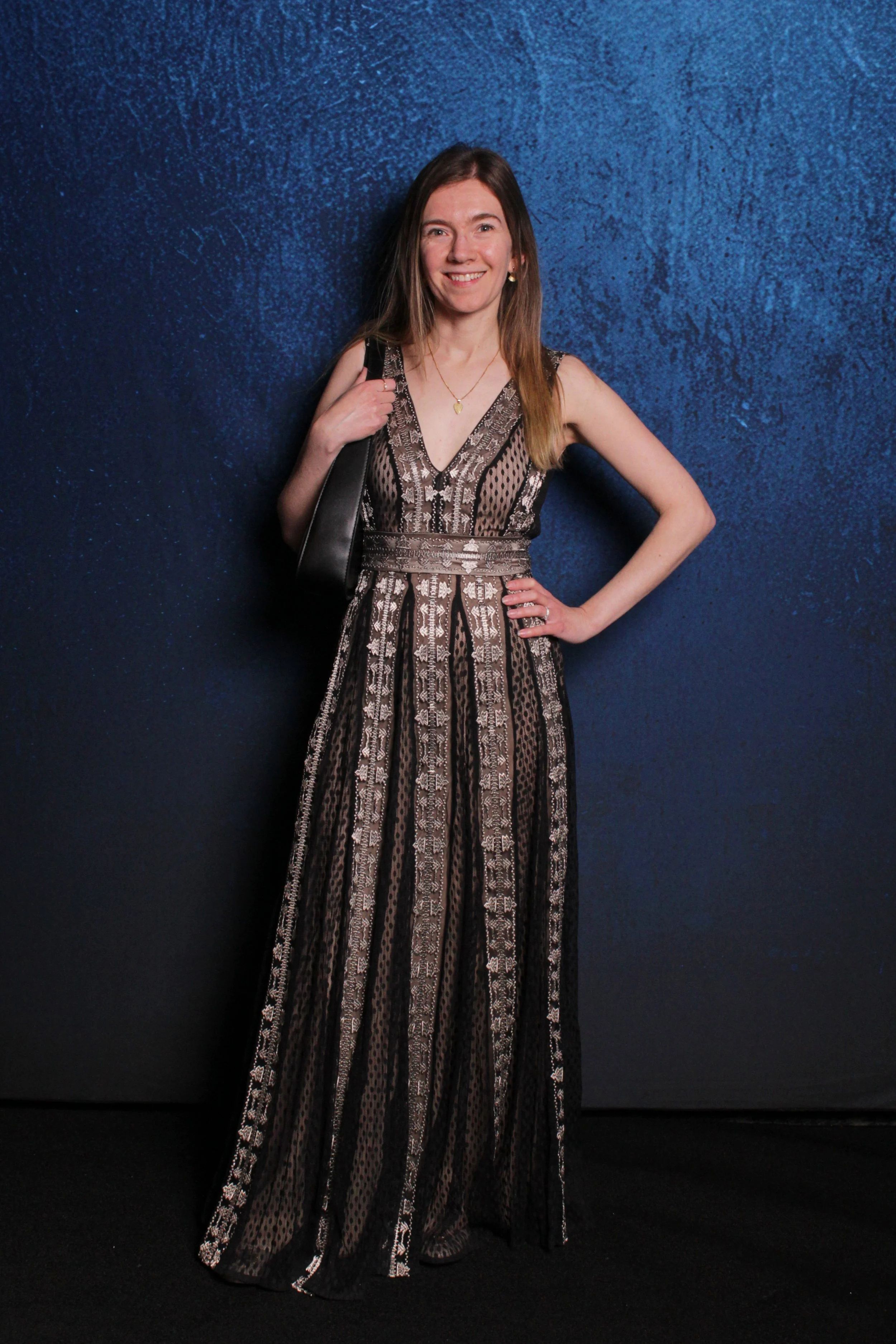 Woman in a patterned full-length dress standing against a blue wall, smiling, with a black handbag on her shoulder.