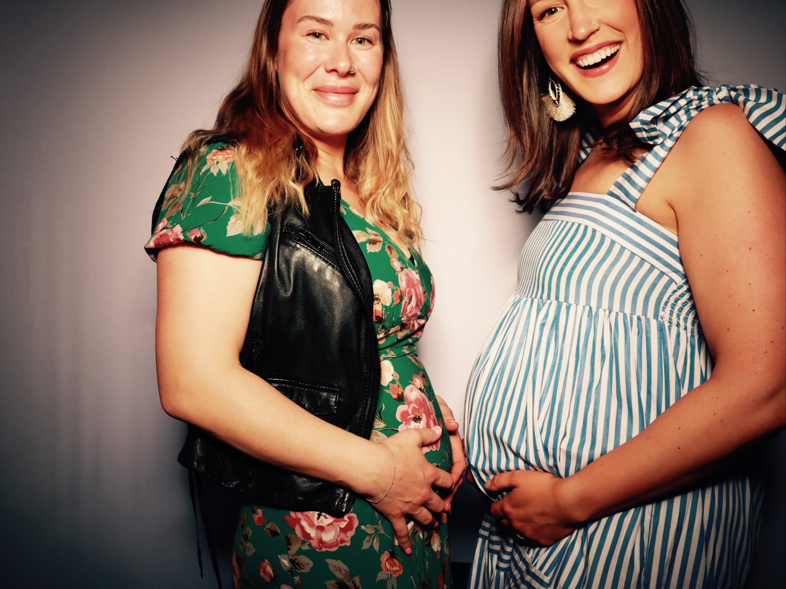 Two women smiling and holding their pregnant bellies, standing close together against a plain background.