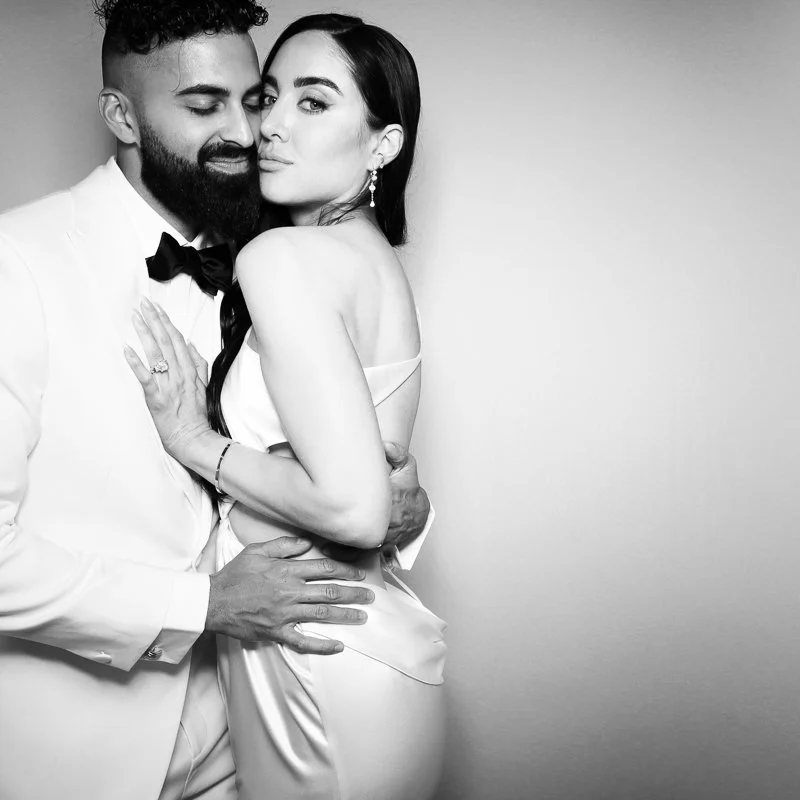 A black and white photograph of a couple dressed formally, embracing each other closely. The man has a beard, is wearing a tuxedo with a bow tie, and the woman has long dark hair, earrings, and a satin dress. They are standing against a plain backgro