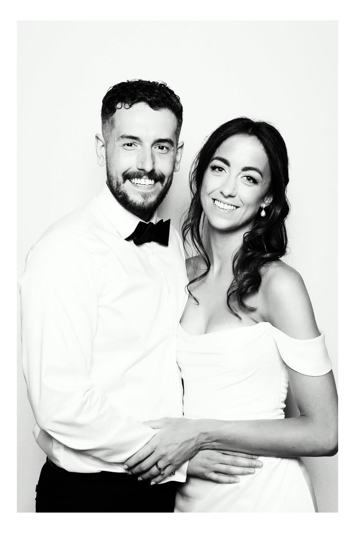 Black and white portrait of a smiling couple dressed in formal attire, with the man wearing a white shirt and bow tie and the woman in a white off-shoulder dress, standing close together against a plain background.