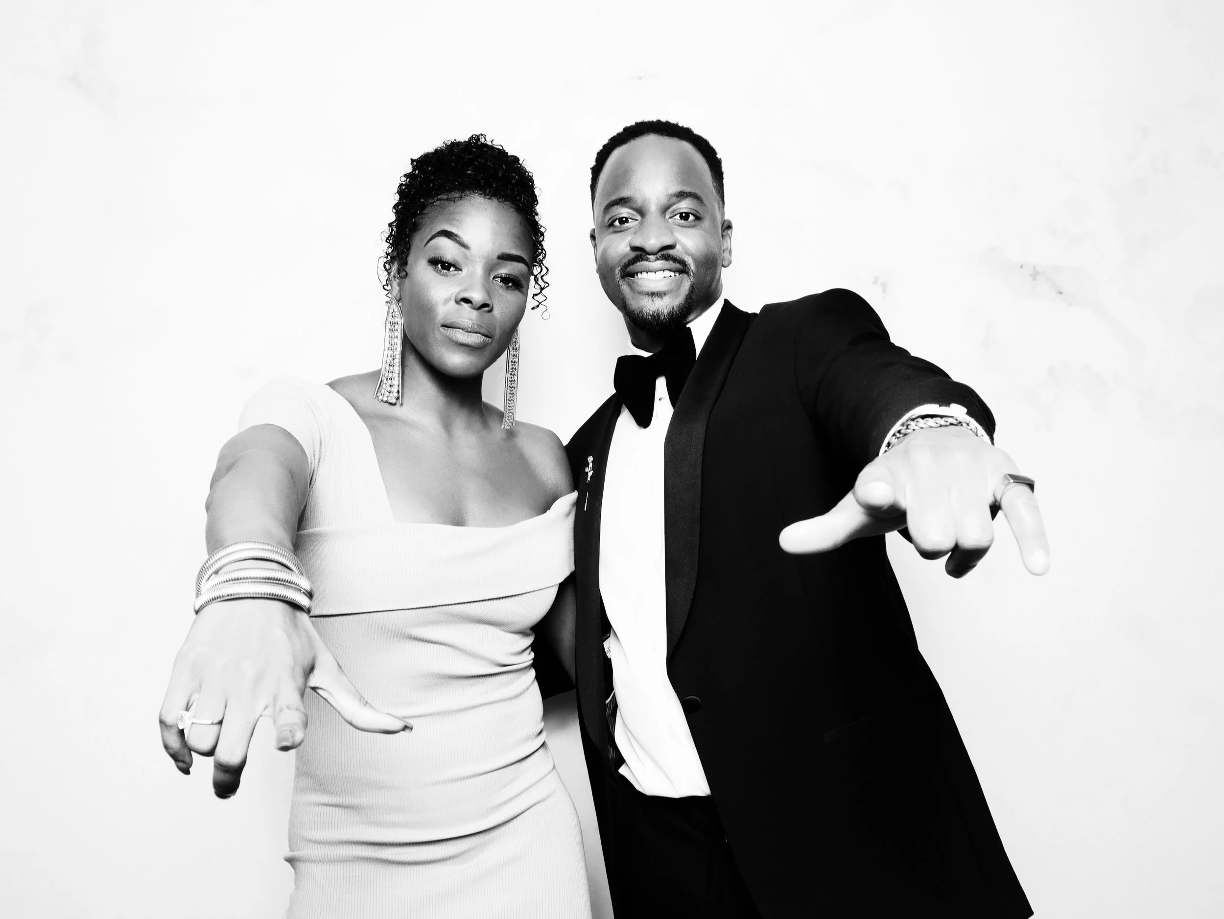 A well-dressed couple posing together in black and white, with both extending their hands towards the camera.