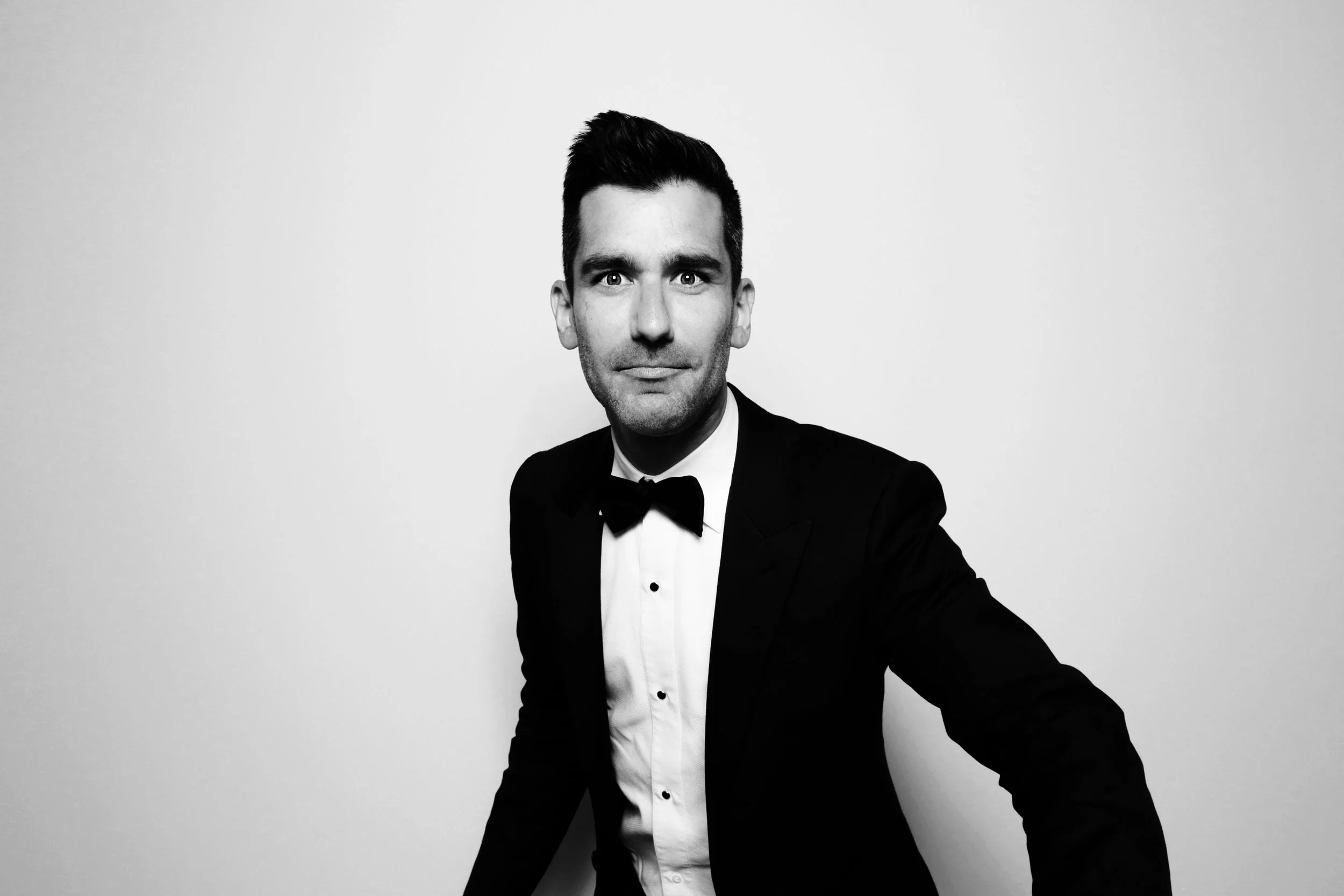 Black and white portrait of a man in a tuxedo with a bow tie, standing against a plain background.