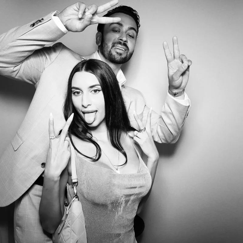 A man and woman posing playfully in front of a plain background, making peace signs and sticking out their tongues, with the woman holding a purse.