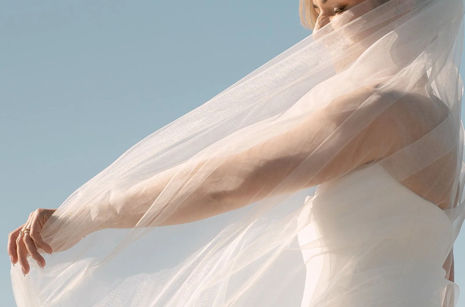 A woman holding a flowing, sheer white fabric in a bright, outdoor setting with a clear blue sky.