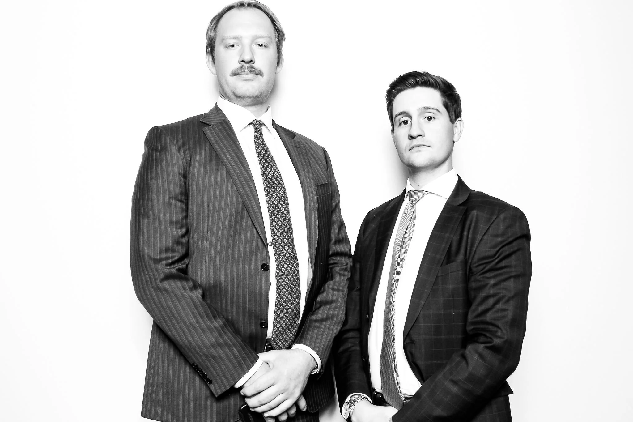 Black and white photo of two men in suits standing against a white background, looking at the camera.