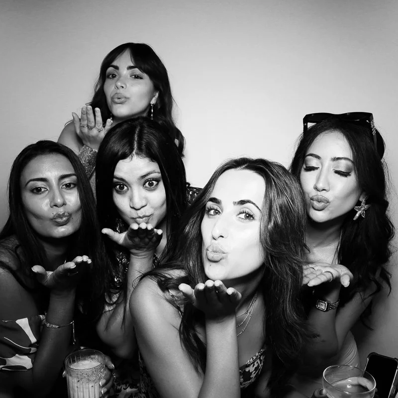 Five women blowing kisses towards the camera in a photo booth, celebrating together