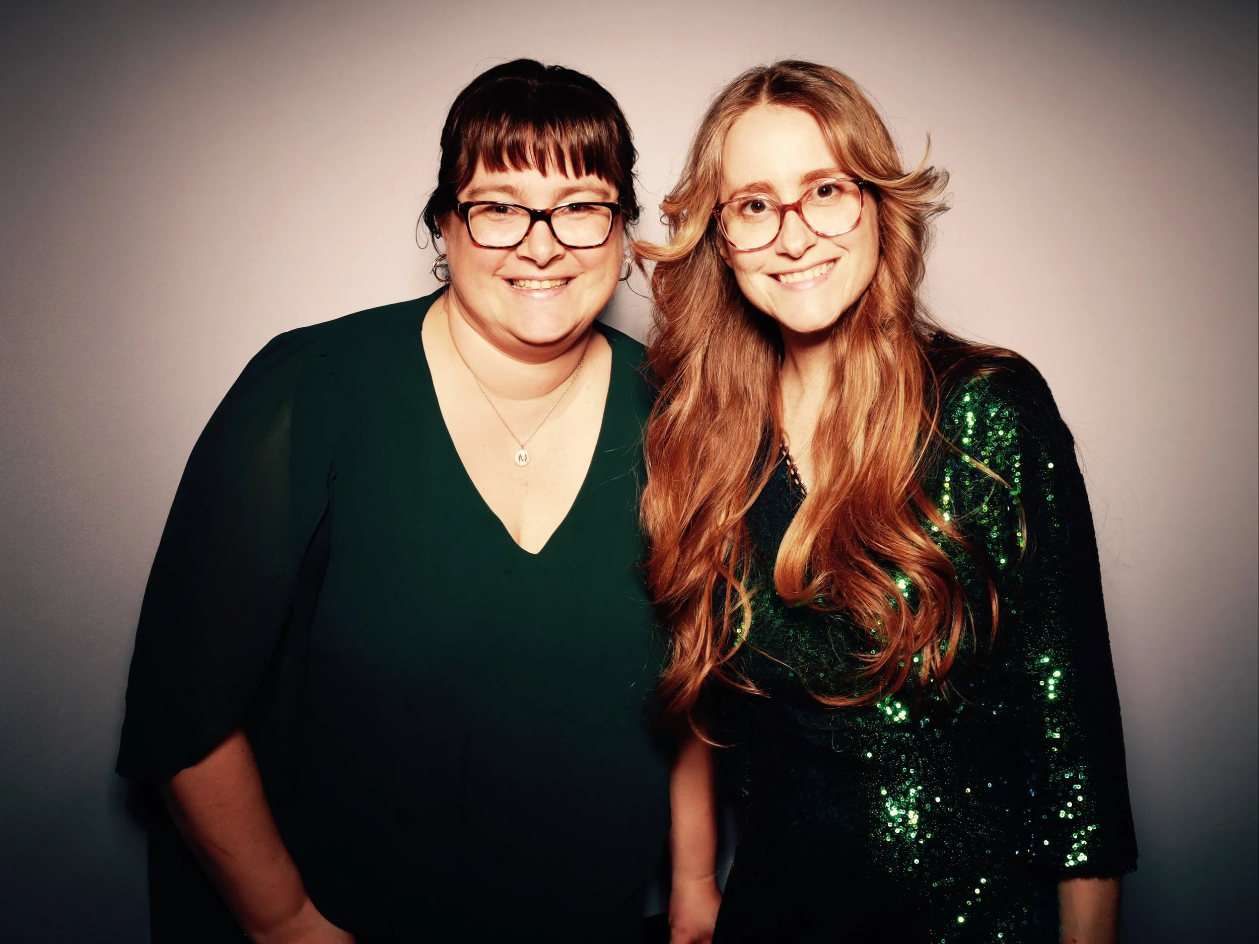 Two women standing side by side, smiling, with a plain gray background. The woman on the left has short dark hair, glasses, and is wearing a dark green top. The woman on the right has long, wavy red hair, glasses, and is wearing a black and green seq