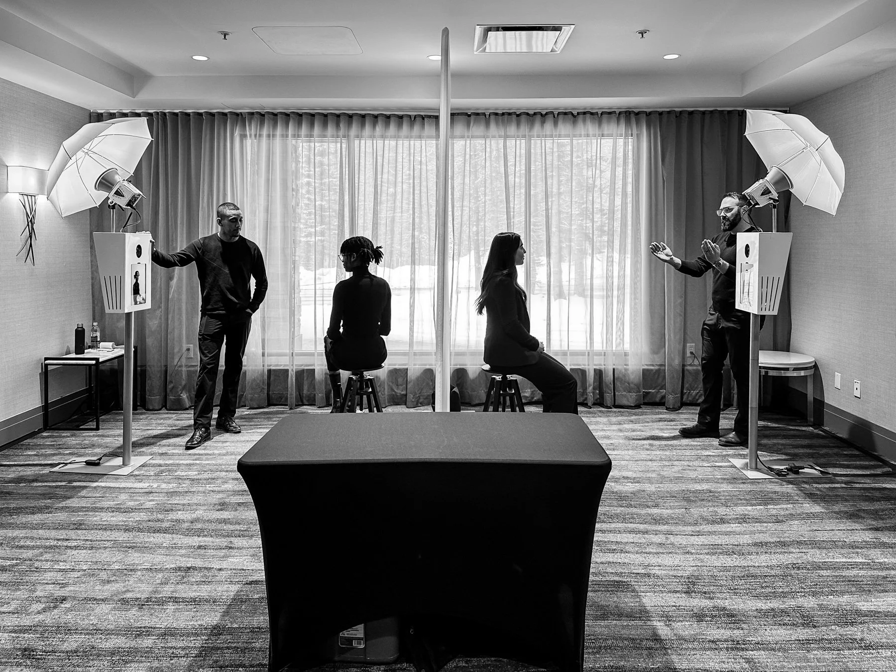 In a studio, two women are seated on stools facing each other, with two men standing on either side. The men are setting up lighting equipment with umbrellas, and the women are preparing for a photo shoot, separated by a partition, with a window and 