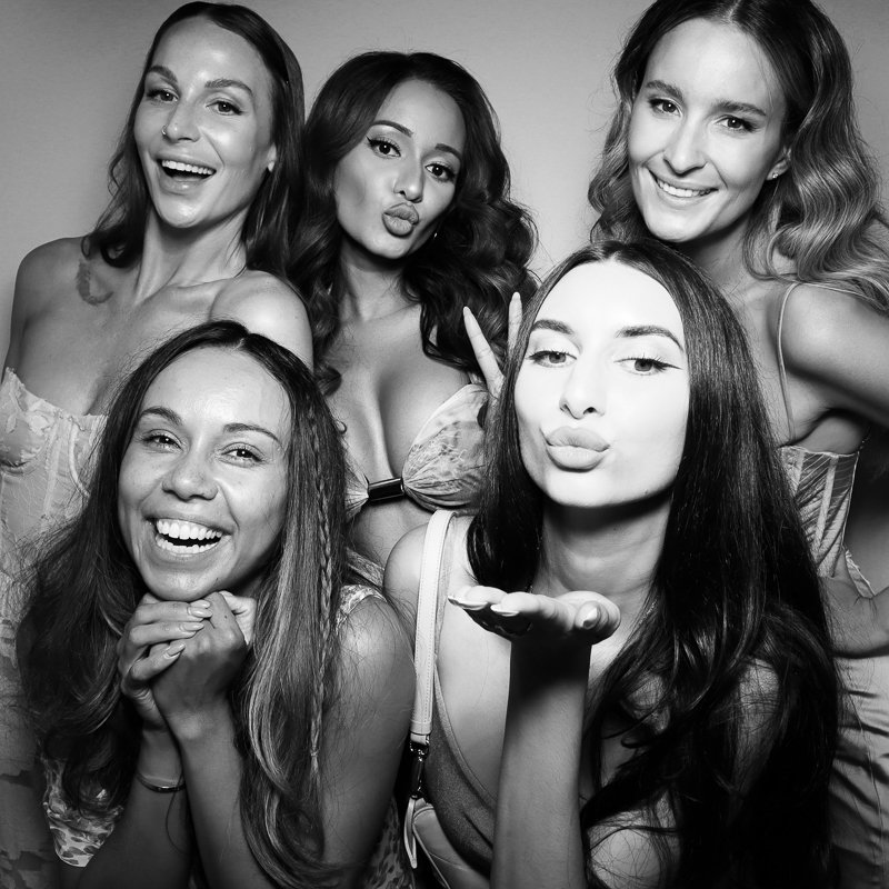 Five women posing together, some smiling and making playful expressions, one blowing a kiss, in a black-and-white photo.