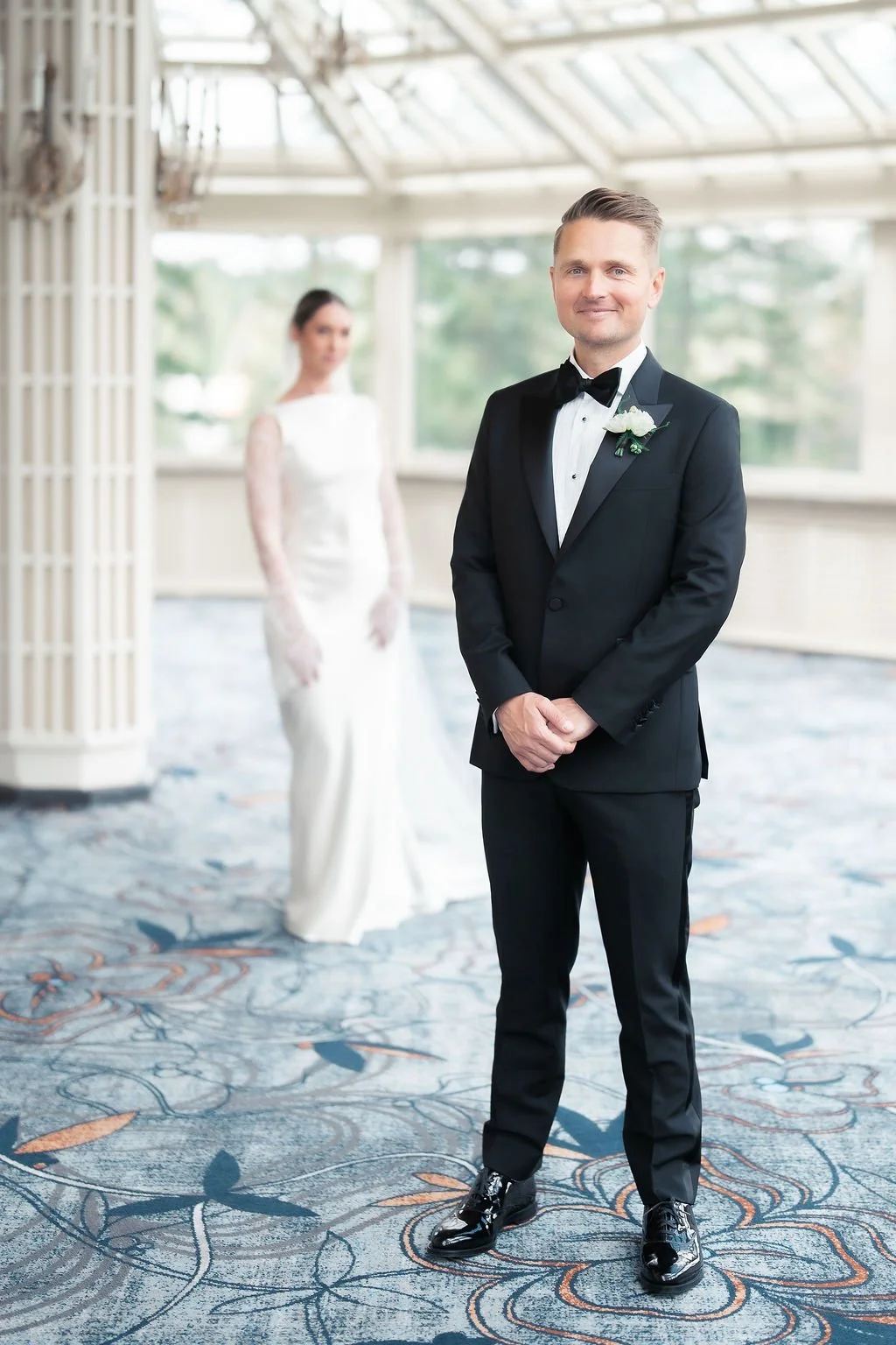 A groom in a black tuxedo with a white boutonniere standing in the foreground, smiling, with a bride in a white wedding dress blurred in the background inside a well-lit venue with large windows and patterned carpet.