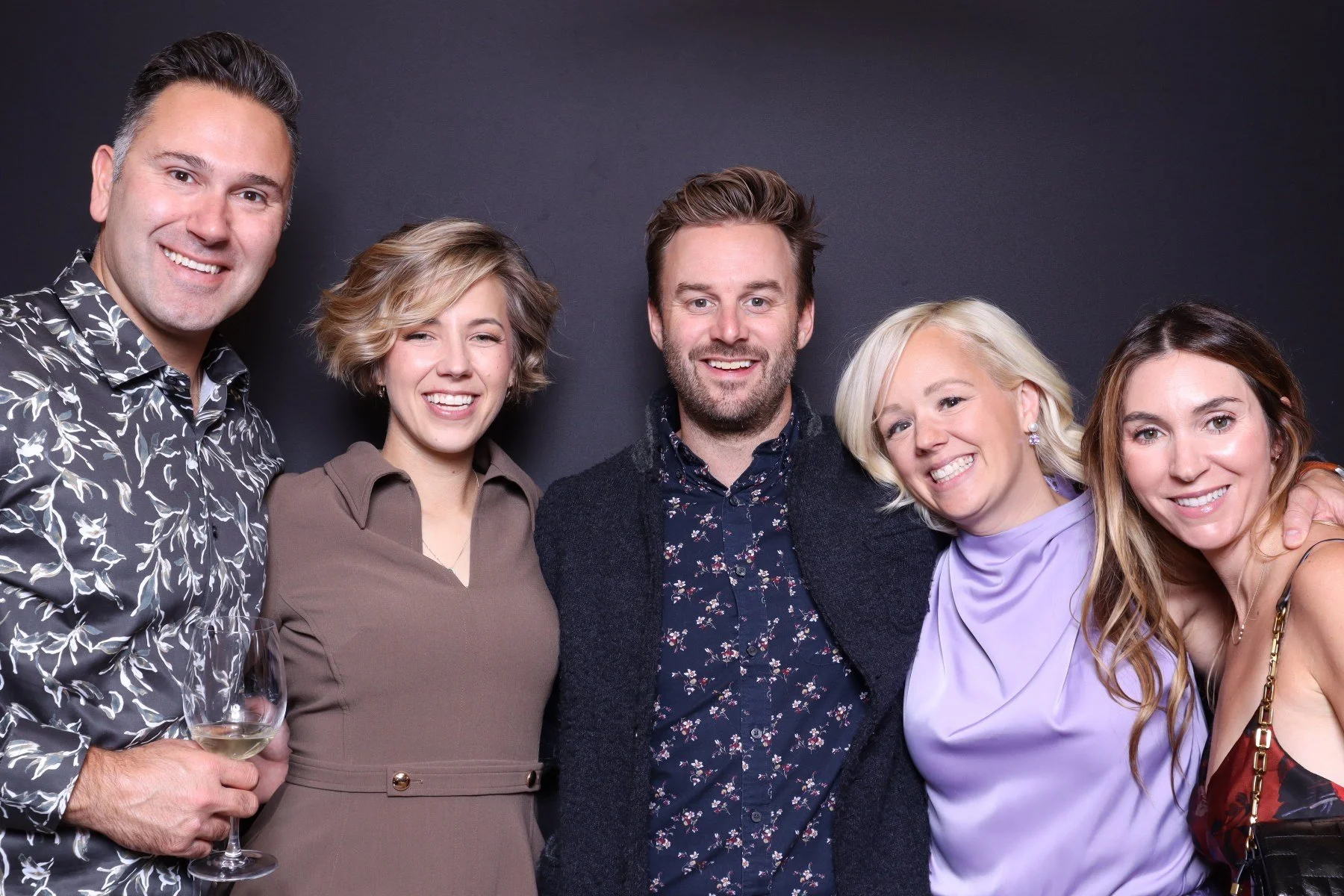 Group of five friends standing close together, smiling, and posing for a photo against a dark background. The person on the far left is holding a glass of white wine.