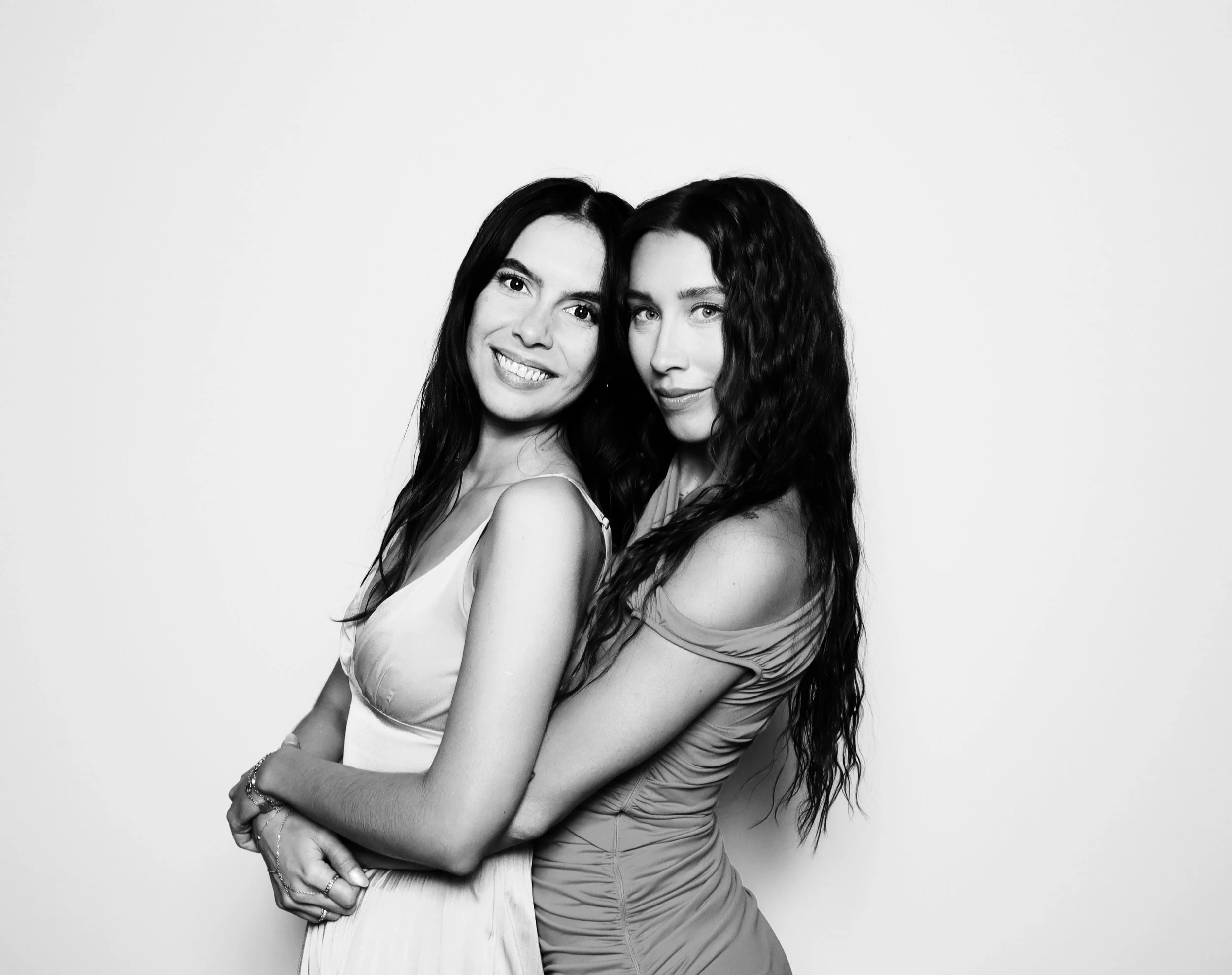 Two women with long, curly and straight dark hair, standing closely together, smiling and looking at the camera, against a plain white background.