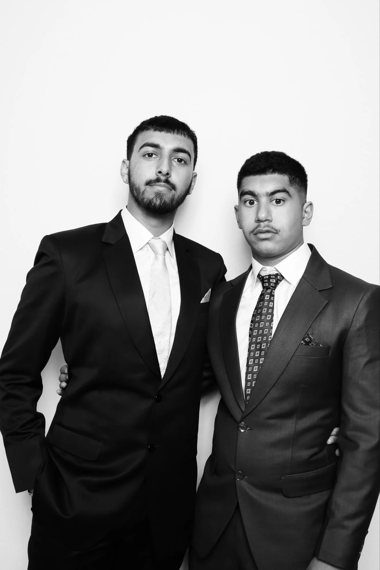 Two men in formal suits standing close together against a plain white wall. The man on the left has a beard and short hair, wearing a dark suit with a light-colored tie. The man on the right has short hair and a light mustache, wearing a dark suit wi