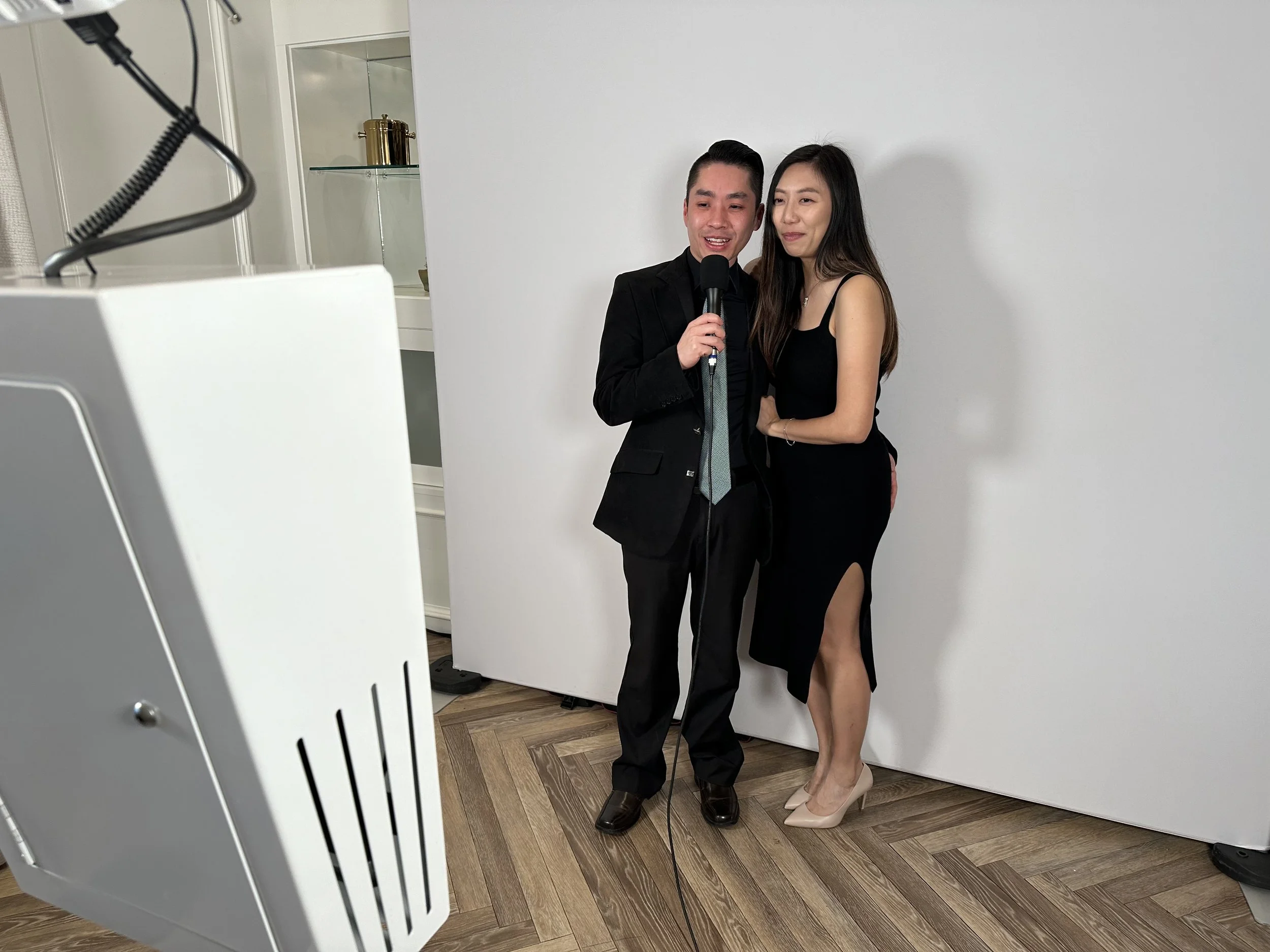 A man and woman are taking a selfie together in front of a white backdrop. The man is holding a microphone and dressed in a black suit, while the woman is wearing a black dress with a slit and beige heels.
