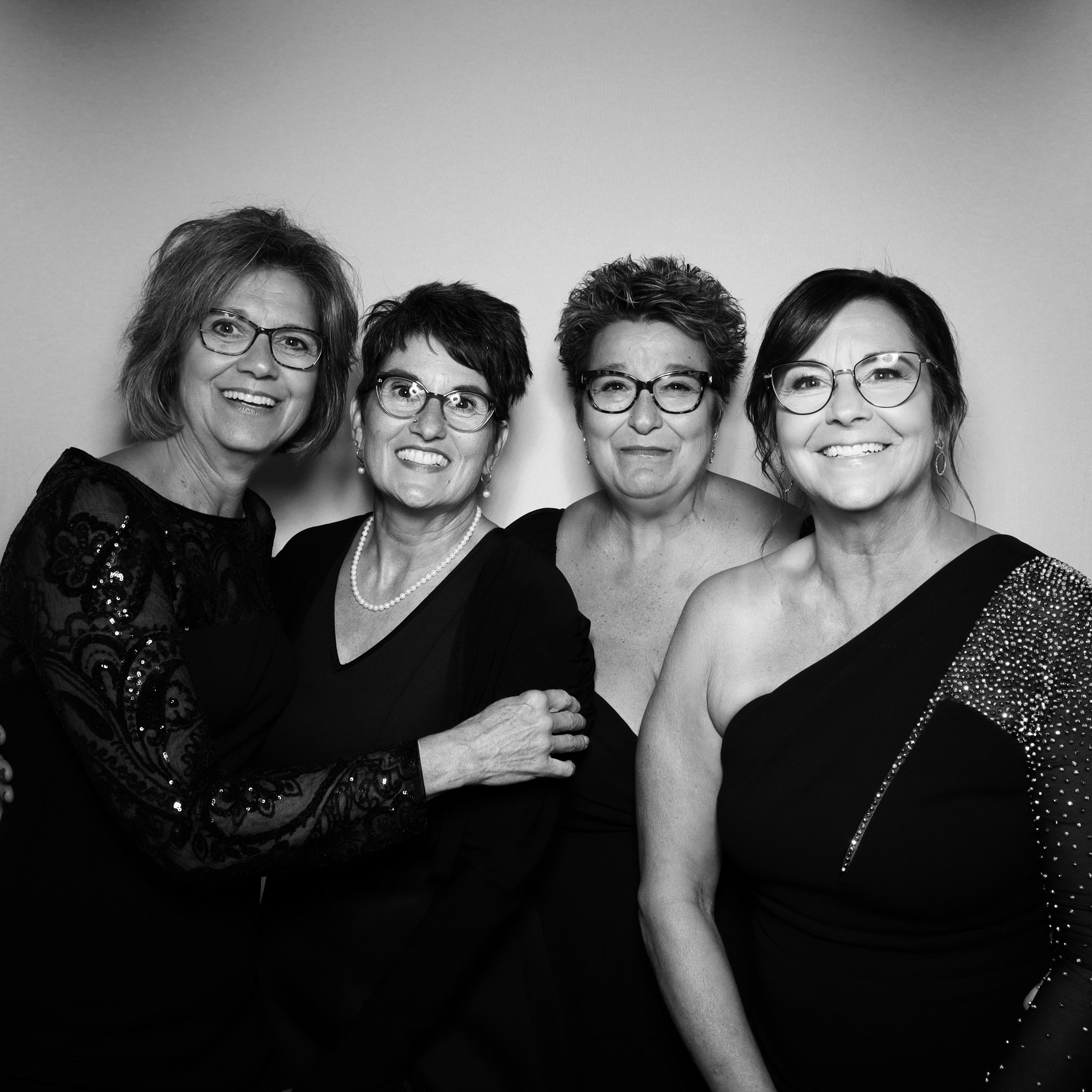 Black and white photo of four women wearing elegant dresses and glasses, standing close together and smiling.