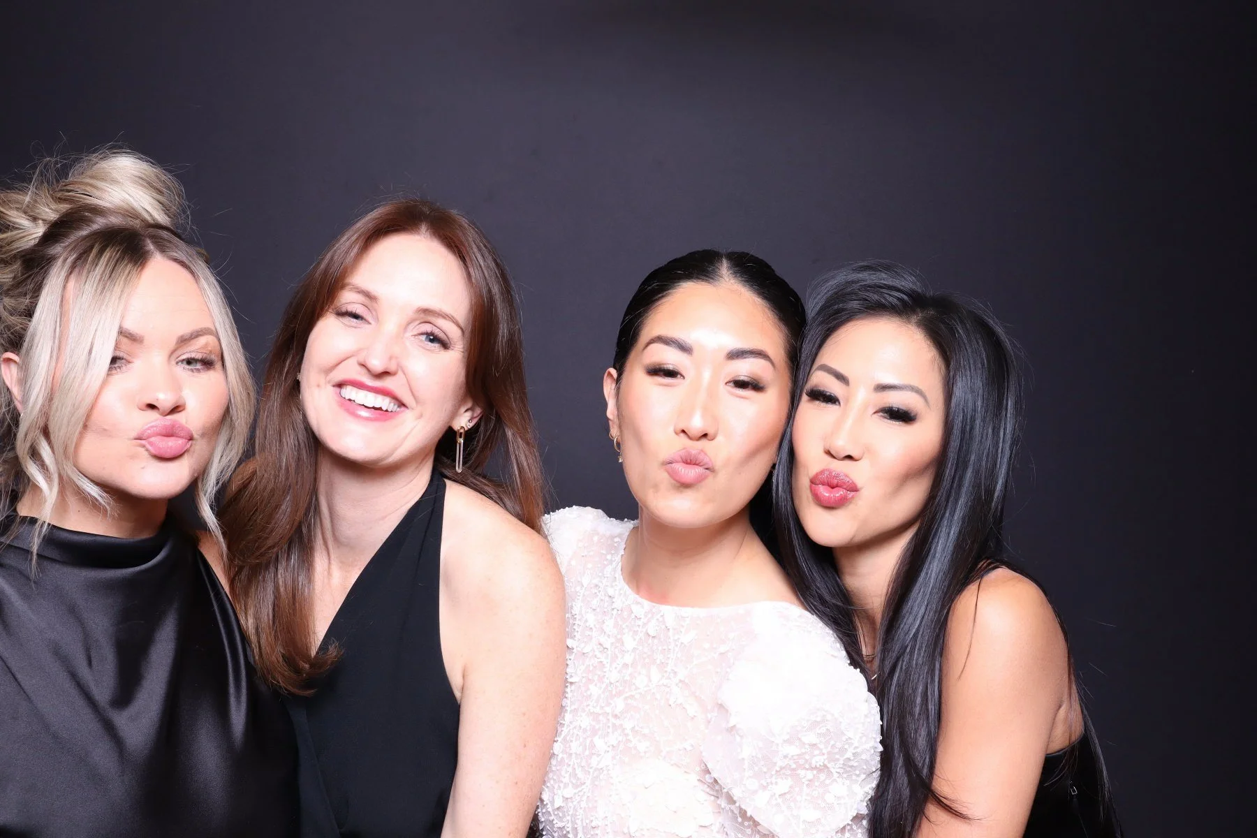 Four women standing close together, making kissy faces, against a dark background.