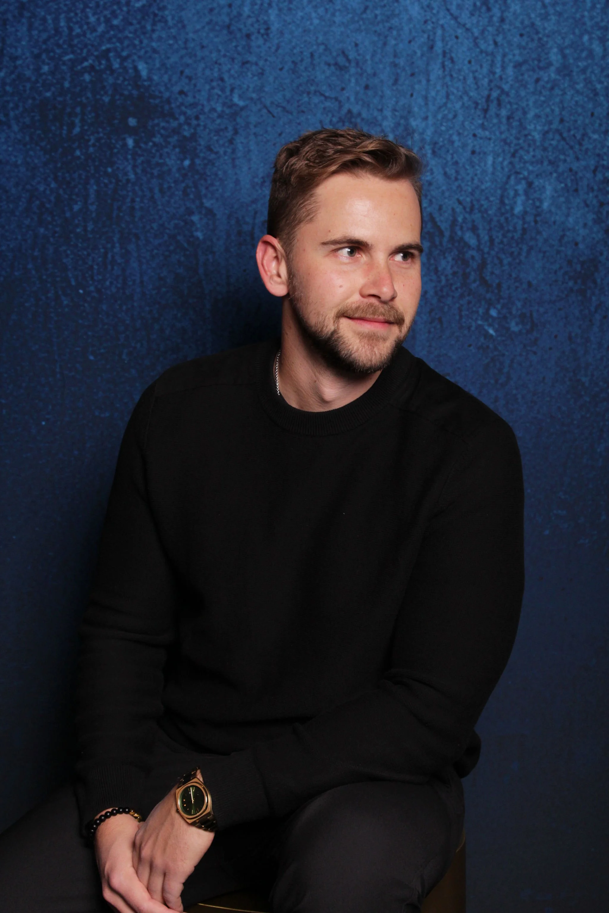 A man with light brown hair and a beard sitting against a textured blue background, wearing a black sweater and a watch on his left wrist.
