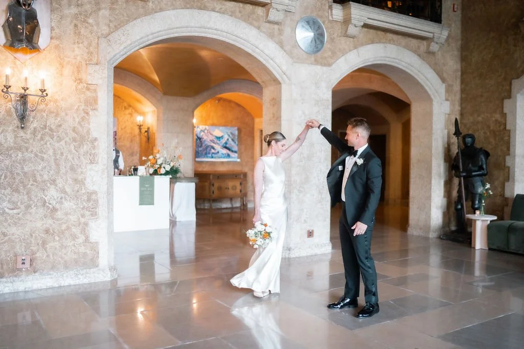 A bride and groom dance together indoors at their wedding reception, with the groom twirling the bride.