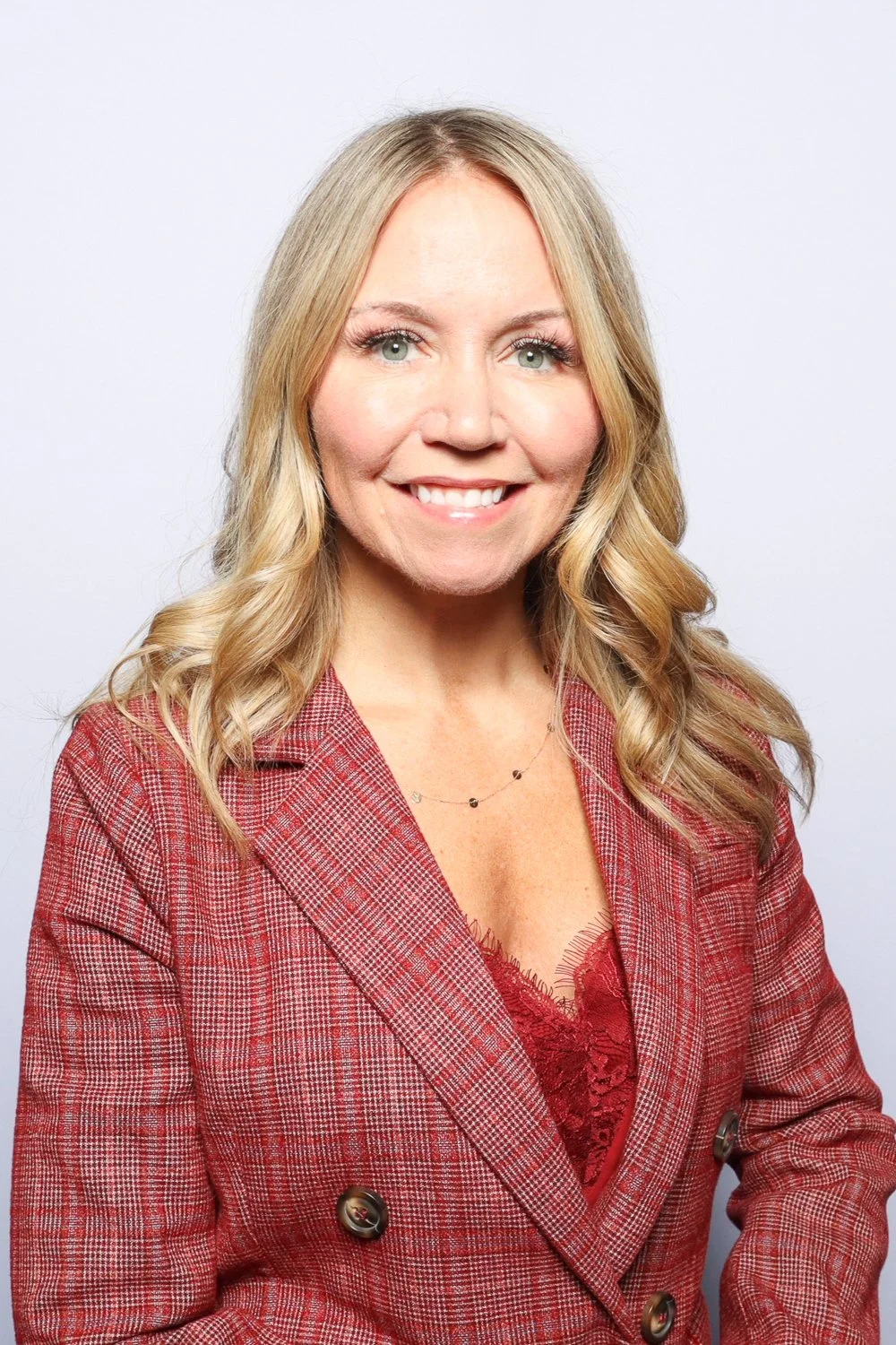 A portrait of a smiling blonde woman with wavy hair, wearing a red checkered blazer over a red lace top, against a plain light background.