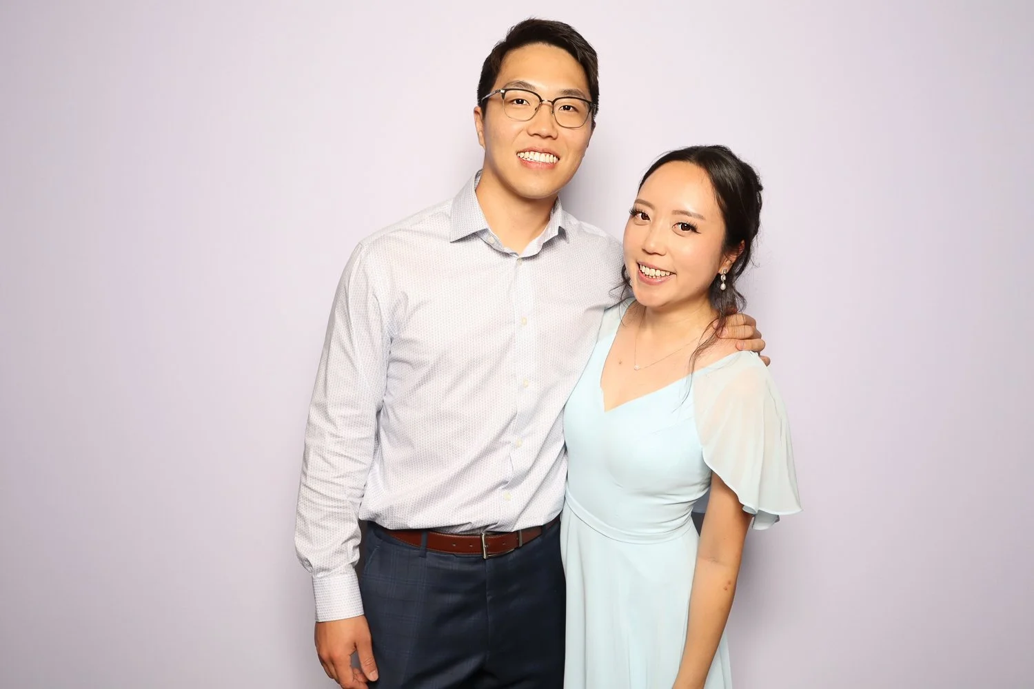 A smiling man and woman standing close together, with the man's arm around the woman's shoulder, against a plain light background.