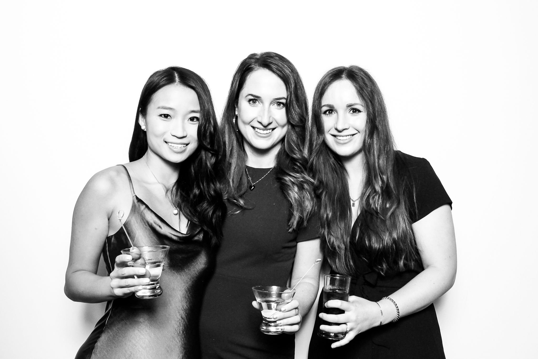 Three women smiling and holding drinks at a social event, standing against a plain white background.